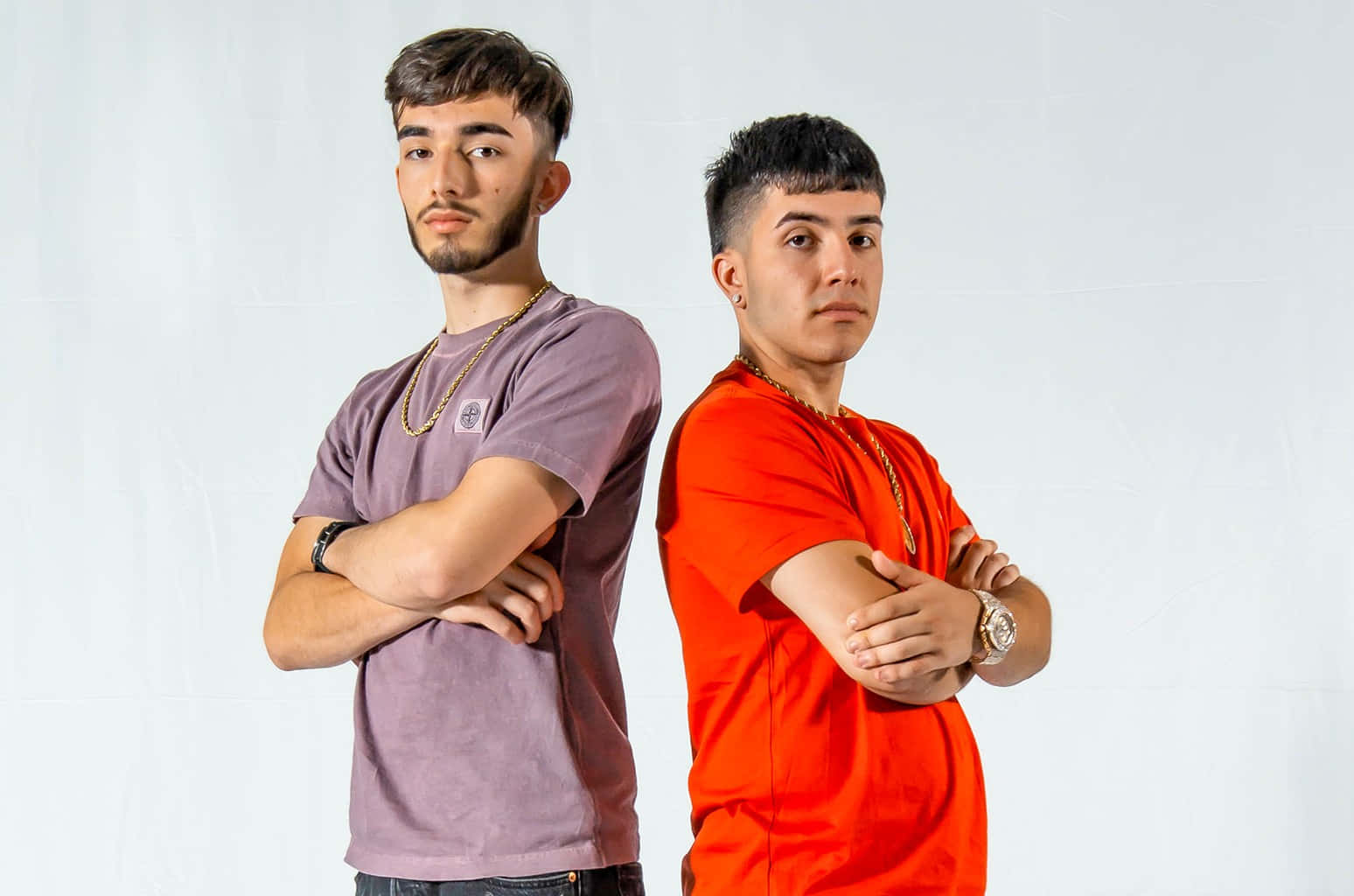 Eslabon Armado 桌布 壁紙 — 兩名年輕男子背對背站立，雙臂交叉，背景是純白色攝影棚，一人穿紫色襯衫，另一人穿亮橙色。