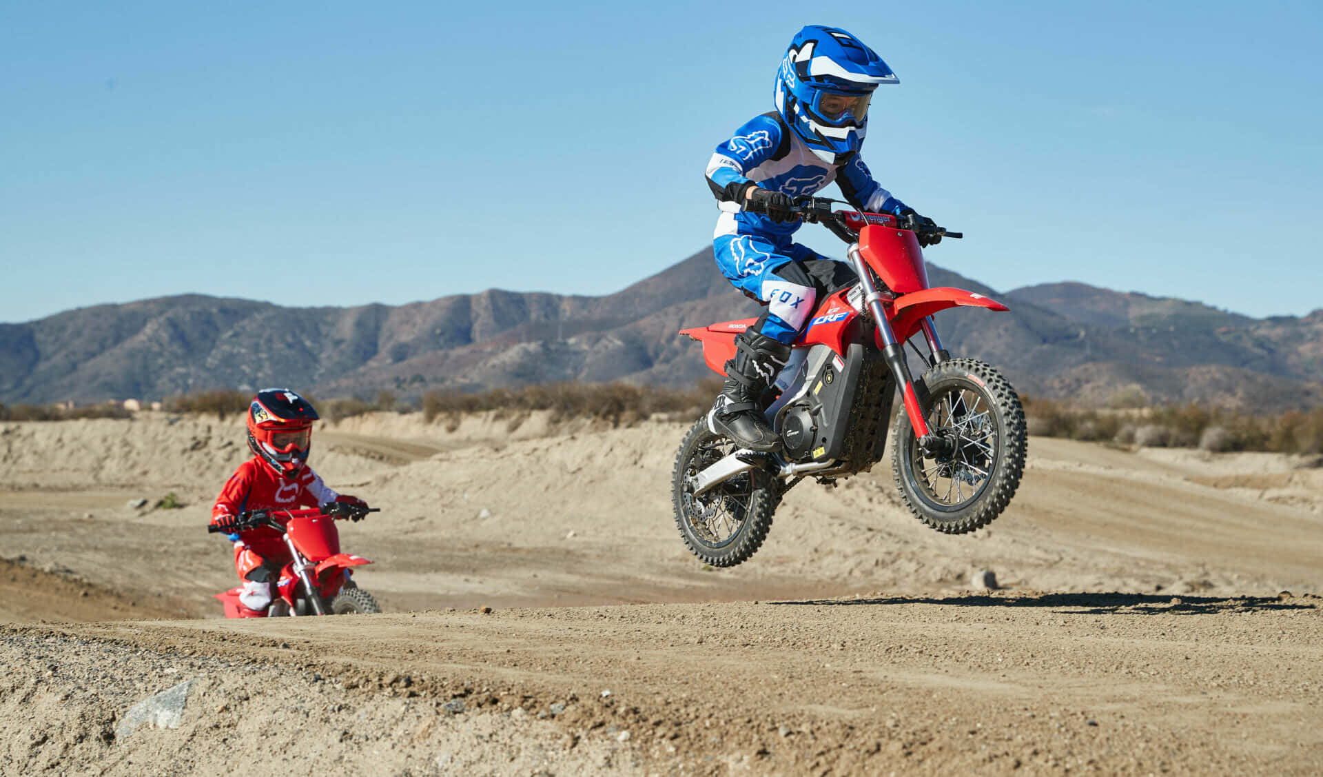 Dirtbike 桌布 壁紙 — 兩名年輕的越野摩托車手全副裝備，在陽光明媚的沙漠賽道上駕駛紅色越野車，背景是山脈。