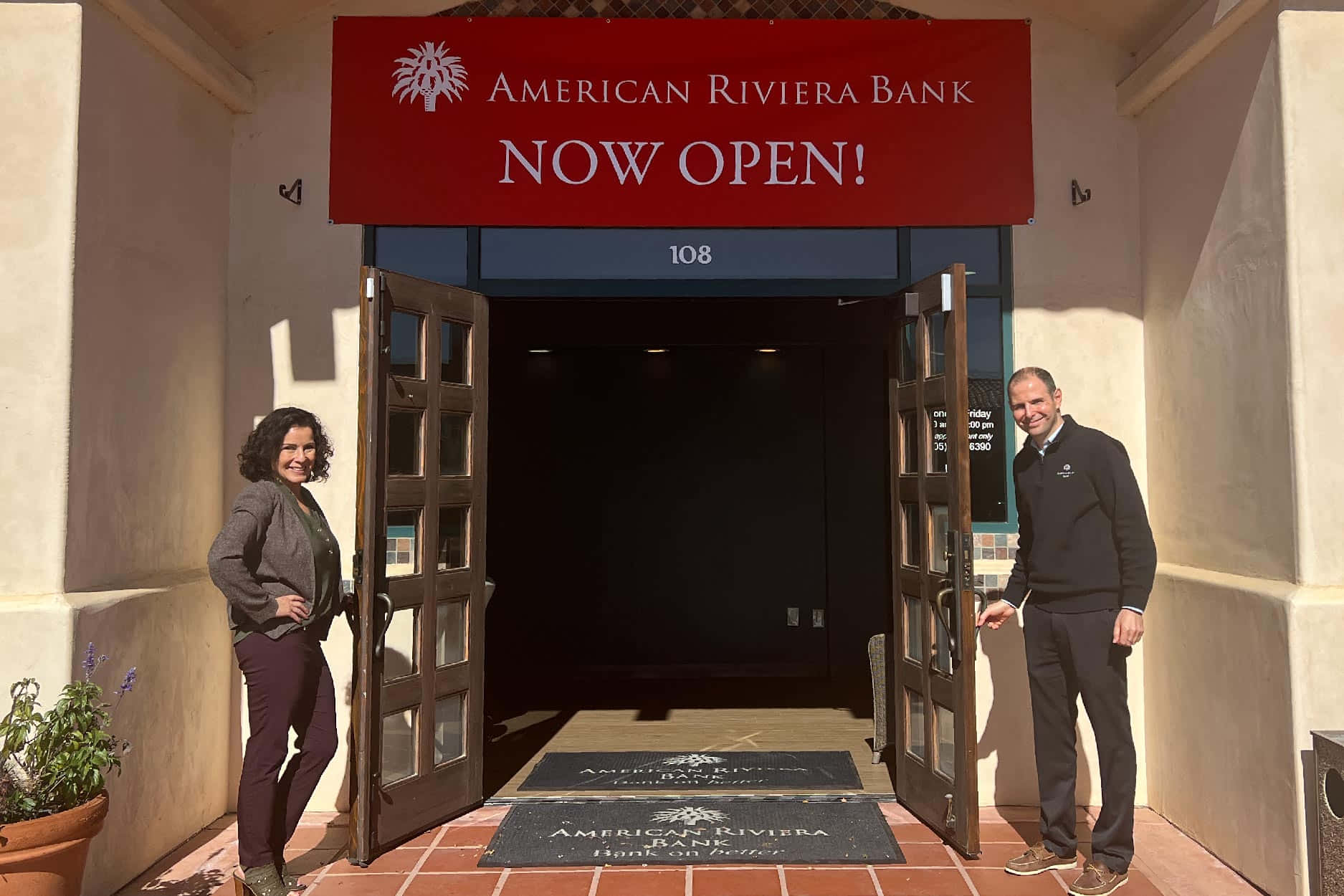 Bank 桌布 壁紙 — 兩名微笑的員工站在新開業的American Riviera Bank敞開的門前，沐浴在明亮的日光下。