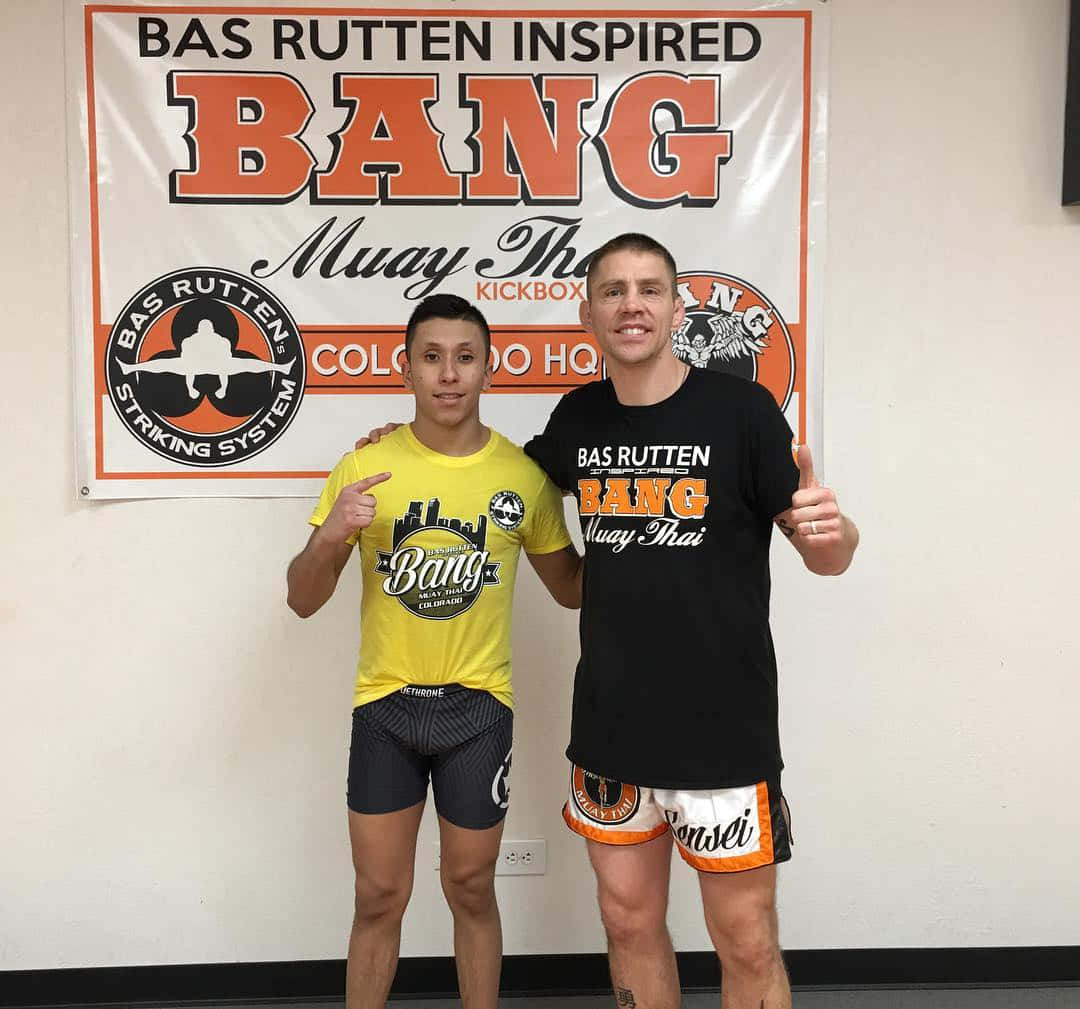 MMA 桌布 壁紙 — 兩名微笑的男士穿著泰拳裝備，在「Bas Rutten Inspired Bang Muay Thai」橫幅前自信擺姿勢。