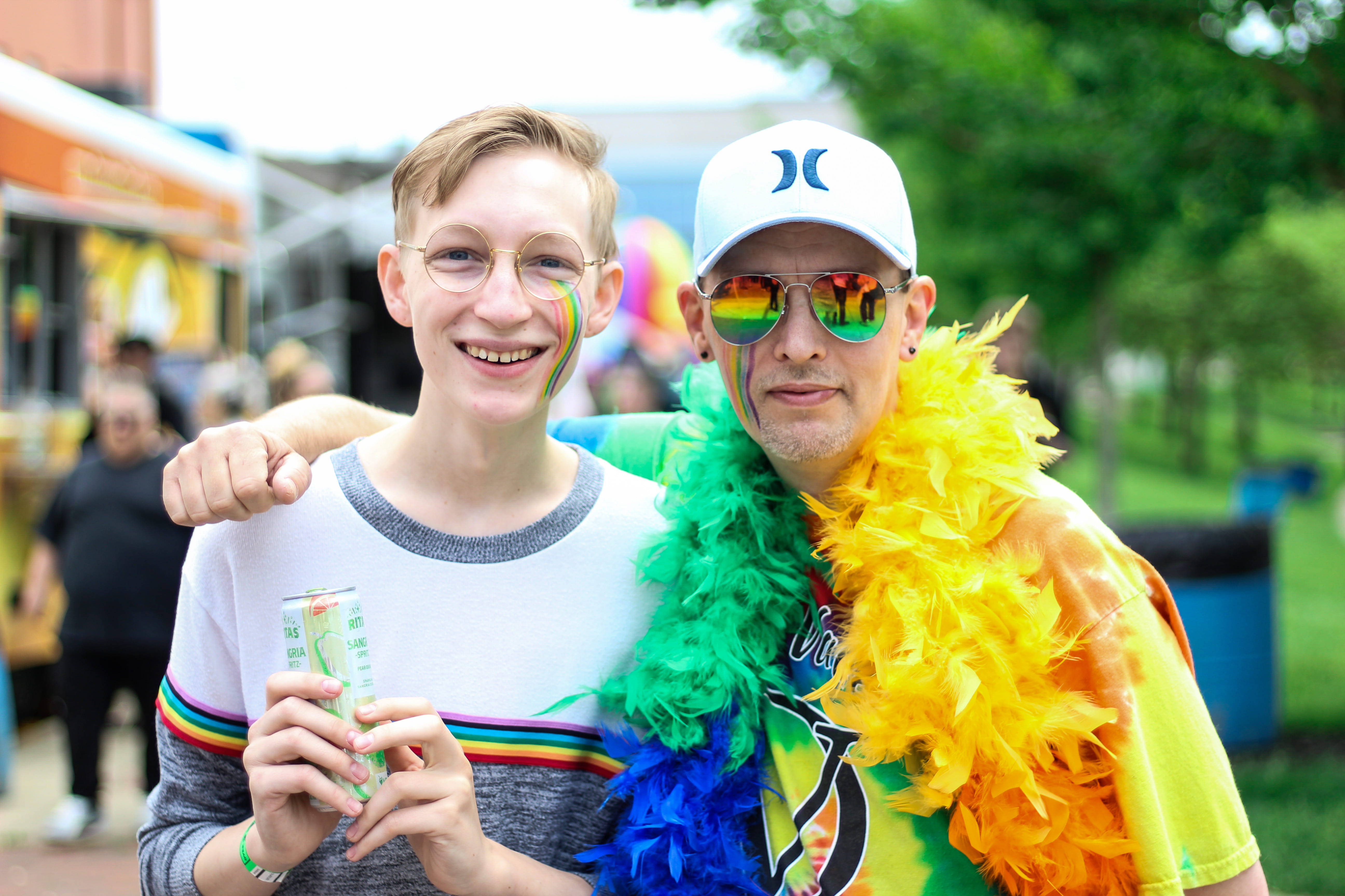 男同志 桌布 壁紙 — 兩名微笑的男子在Pride活動中，臉上塗著彩虹顏料，戴著羽毛圍巾，在戶外拿著飲品。