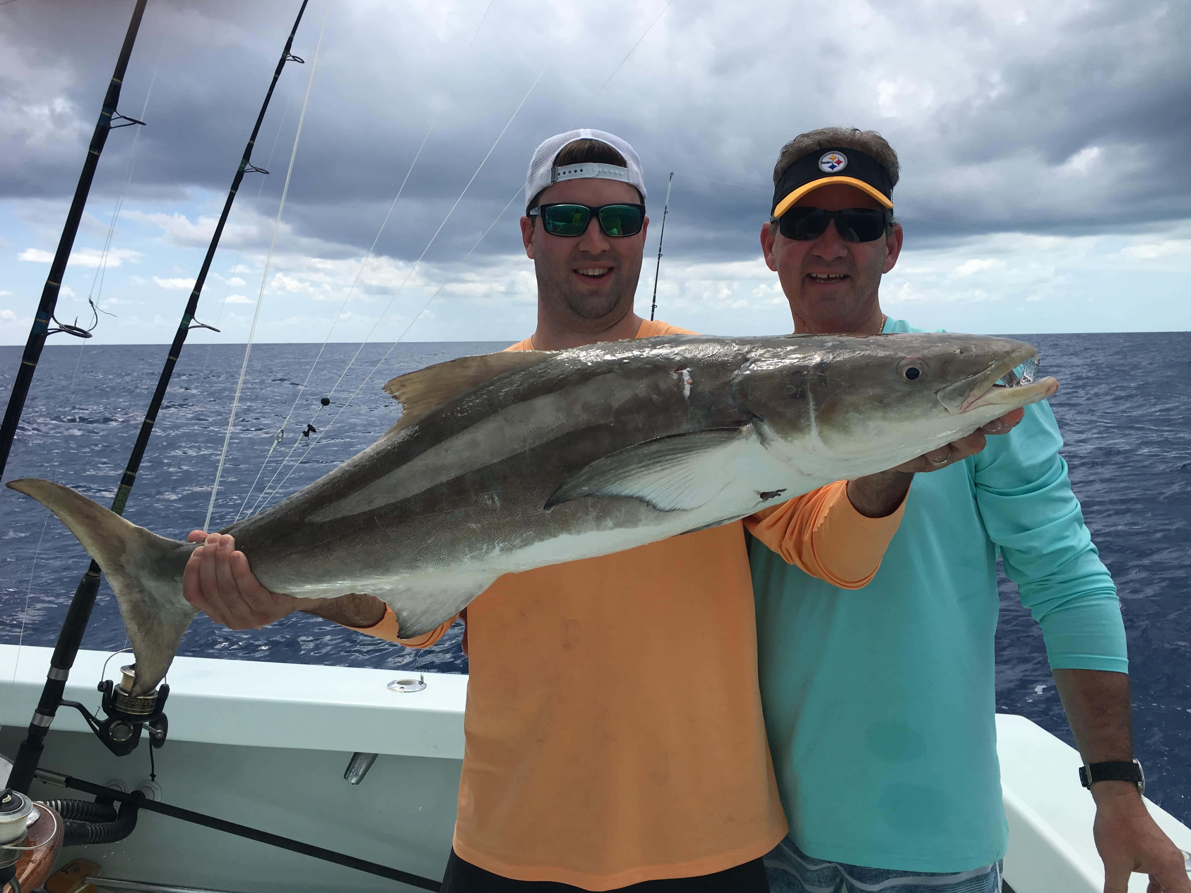 Cobia 桌布 壁紙 — 兩名微笑的男子在船上自豪地舉起一條大銀魚，背景是多雲的海洋。