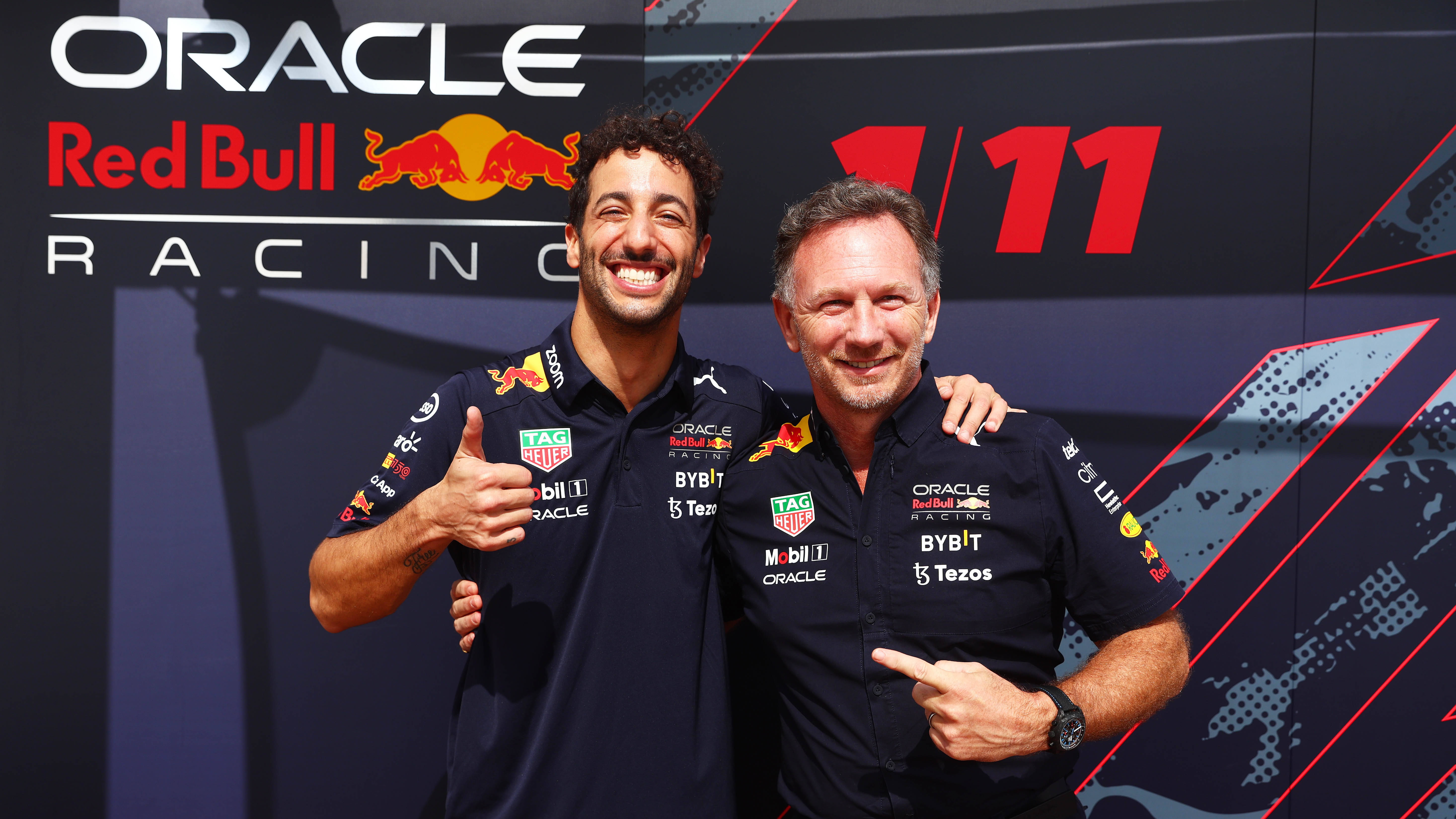 Smile 桌布 壁紙 — 兩名微笑的男子身穿Oracle Red Bull Racing車隊襯衫，在帶有紅色和黑色幾何圖案的品牌背景前愉快地擺姿勢。