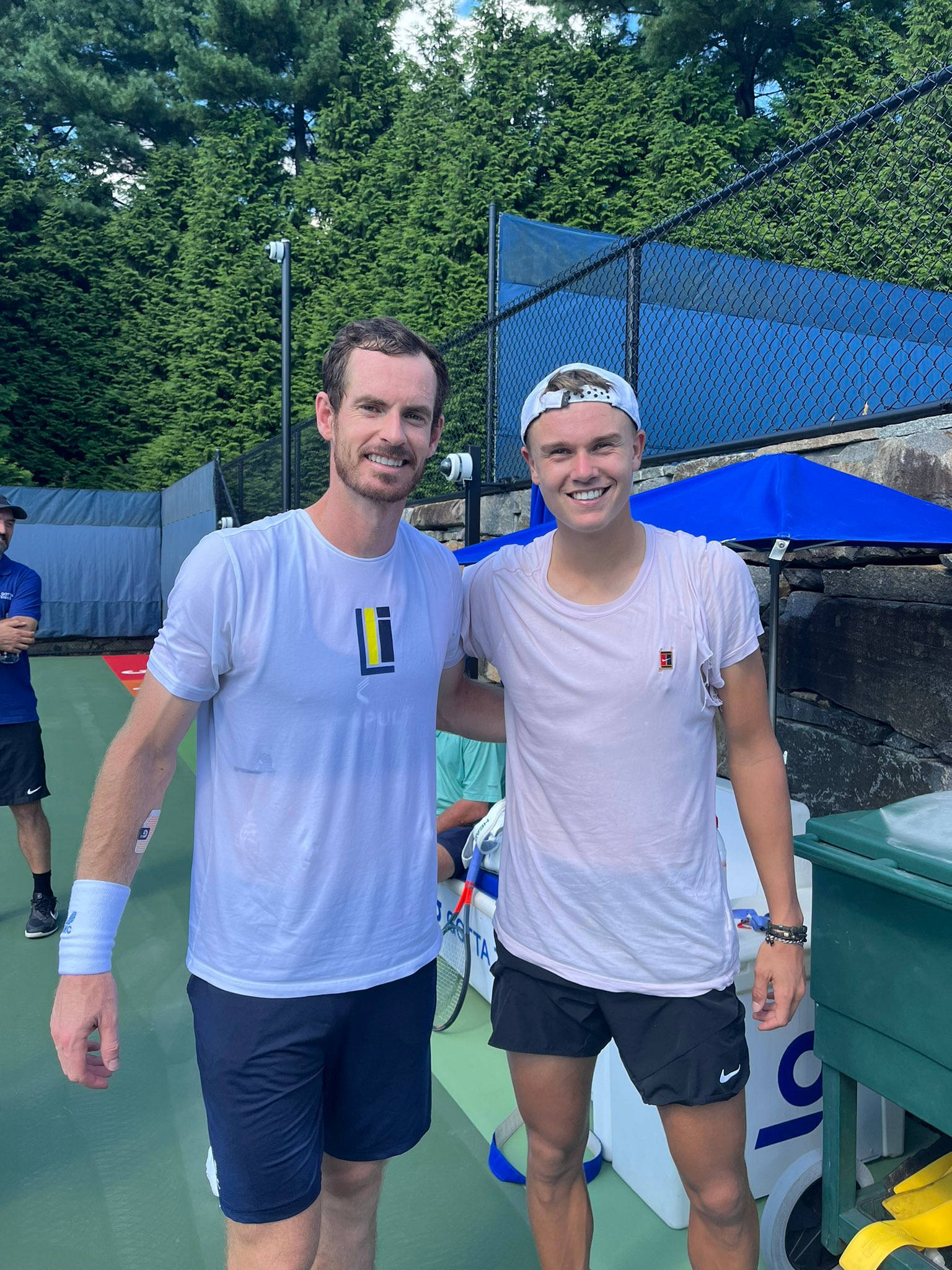 Tennis 桌布 壁紙 — 兩名微笑的男性網球運動員在一個被茂密常綠樹木環繞的綠色球場上一起擺姿勢。
