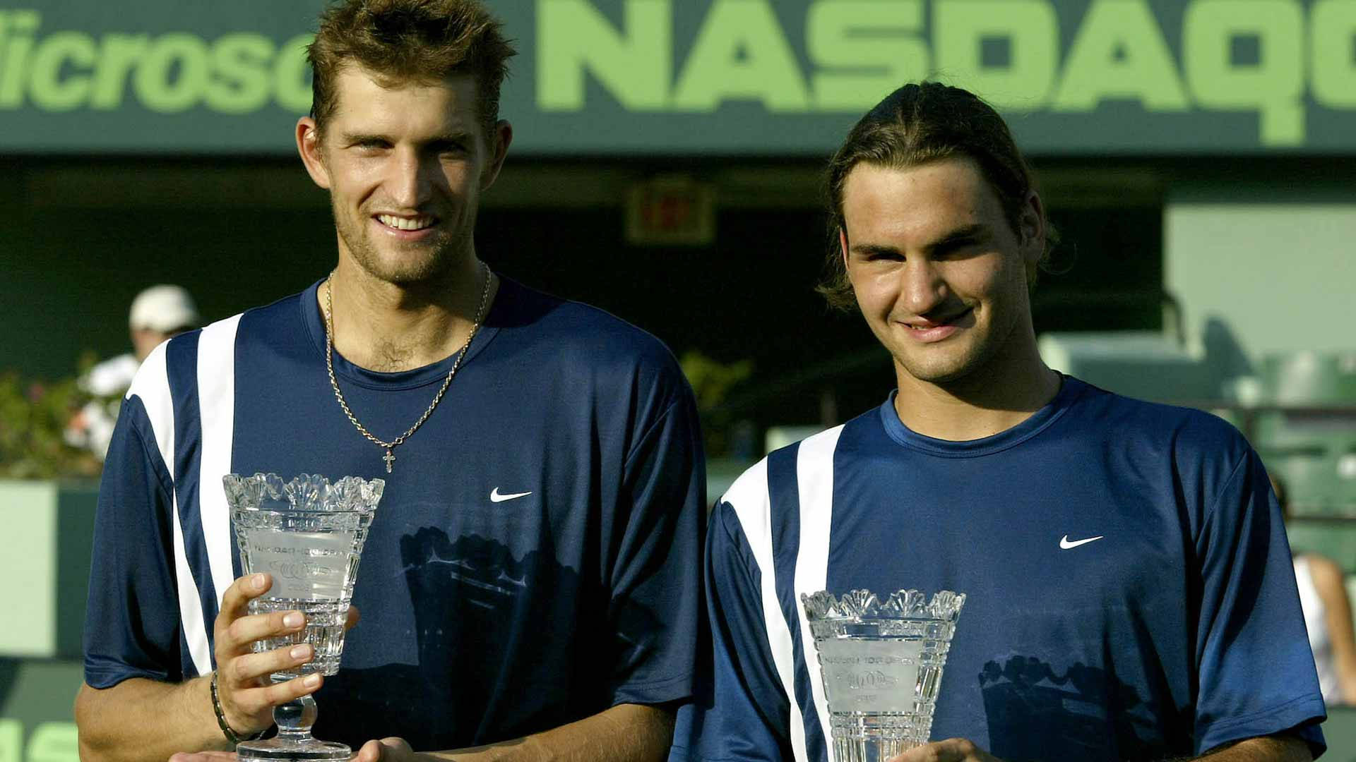 Tennis 桌布 壁紙 — 兩名微笑的男性網球運動員穿著藍色Nike襯衫，在綠色NASDAQ背景下手持透明獎盃。