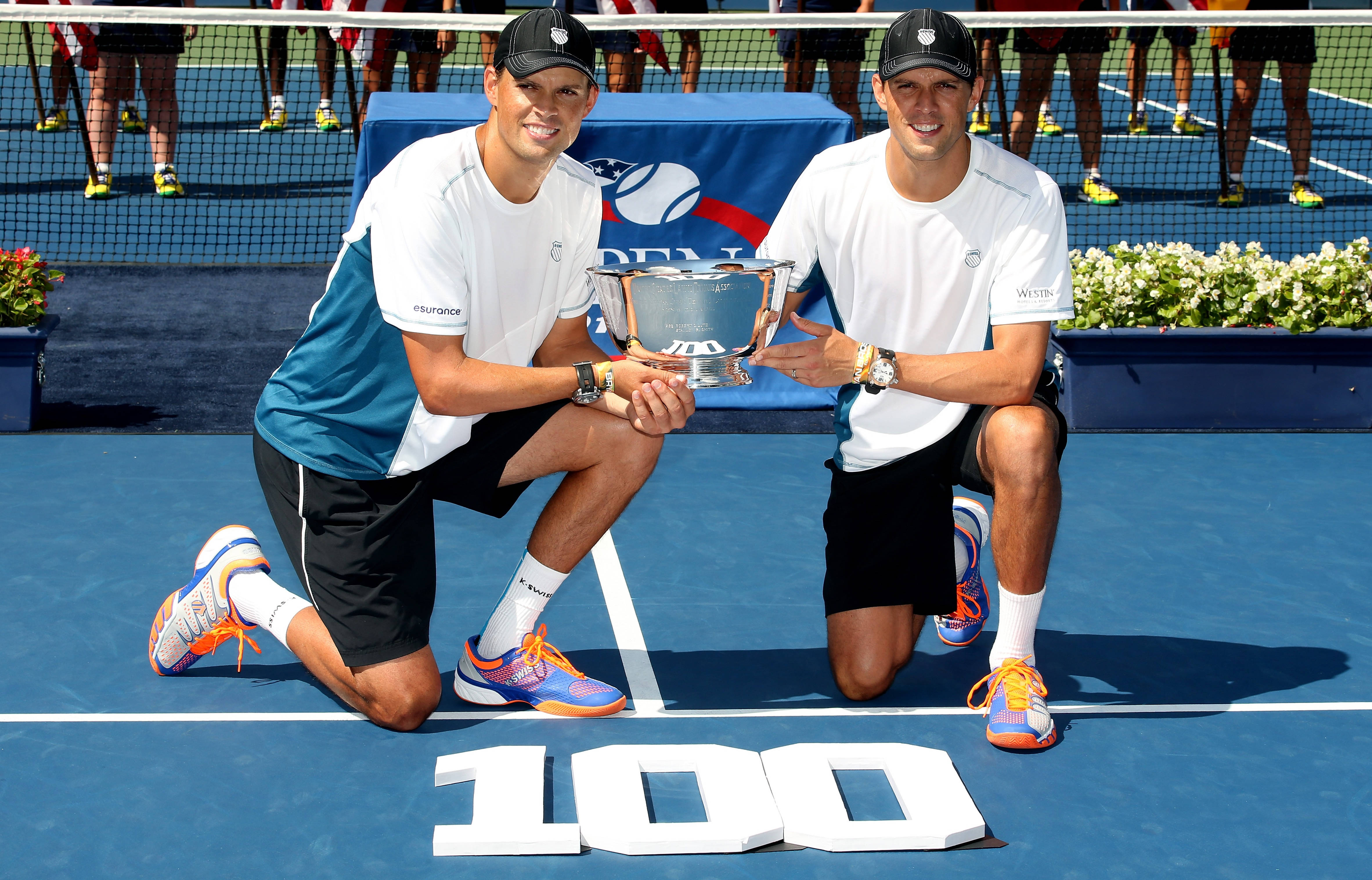Tennis 桌布 壁紙 — 兩名微笑的男性網球選手跪在藍色球場上，手持一個帶有大型白色「100」字母的銀色獎盃。
