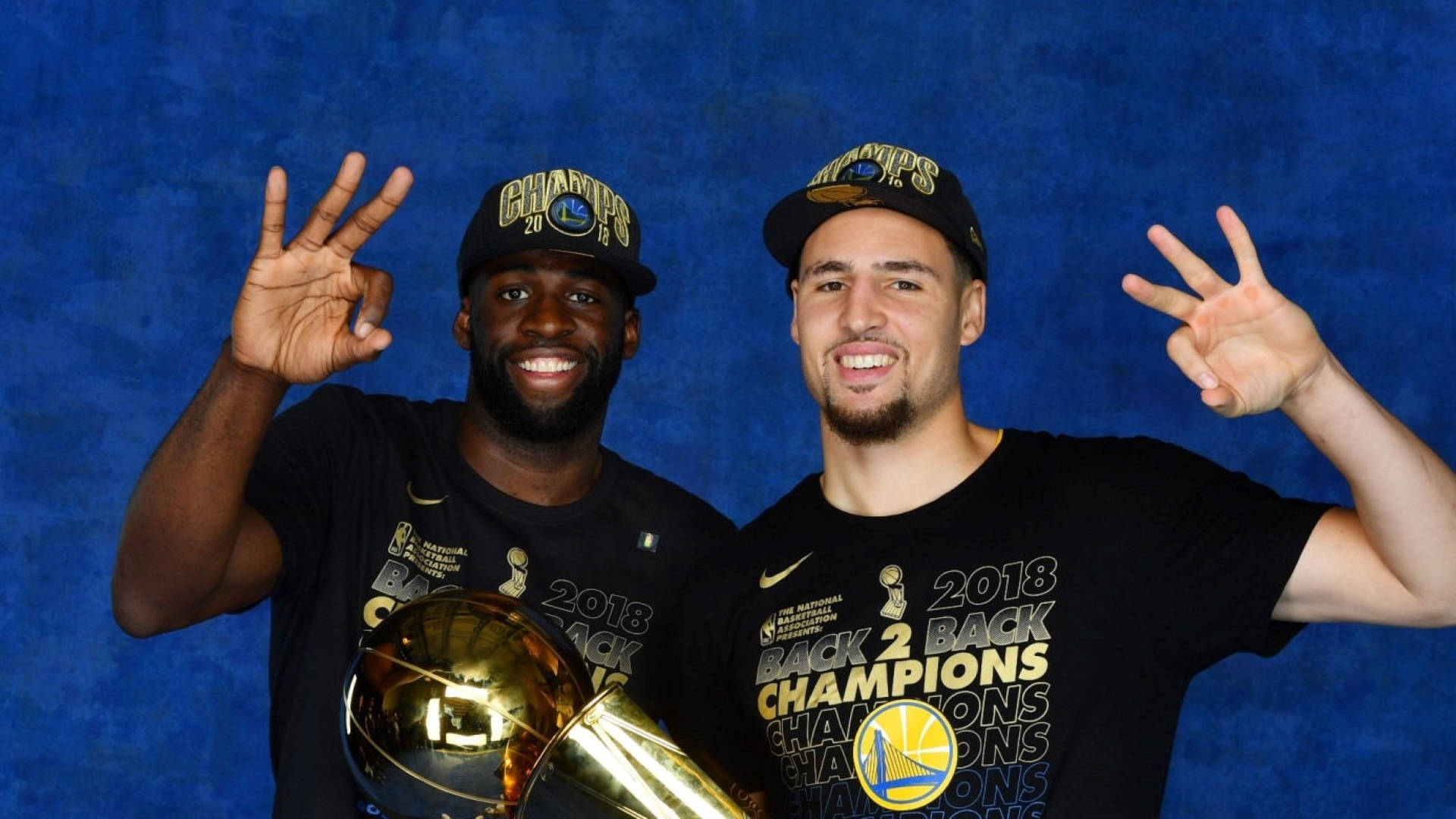 Draymond Green 桌布 壁紙 — 兩名微笑的籃球運動員身穿黑色「2018 Back 2 Back Champions」T恤，在帶有紋理的藍色背景前與金色獎盃合影。
