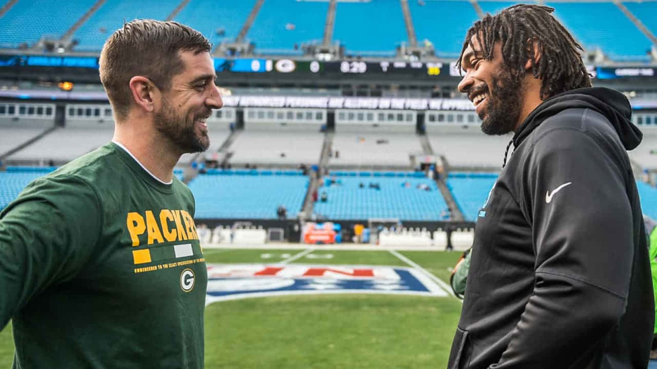 Aaron Rodgers 桌布 壁紙 — 兩名微笑的美式足球員穿著綠色和黑色裝備，在充滿活力的體育場草地上共享時刻，身後是空著的藍色座位。