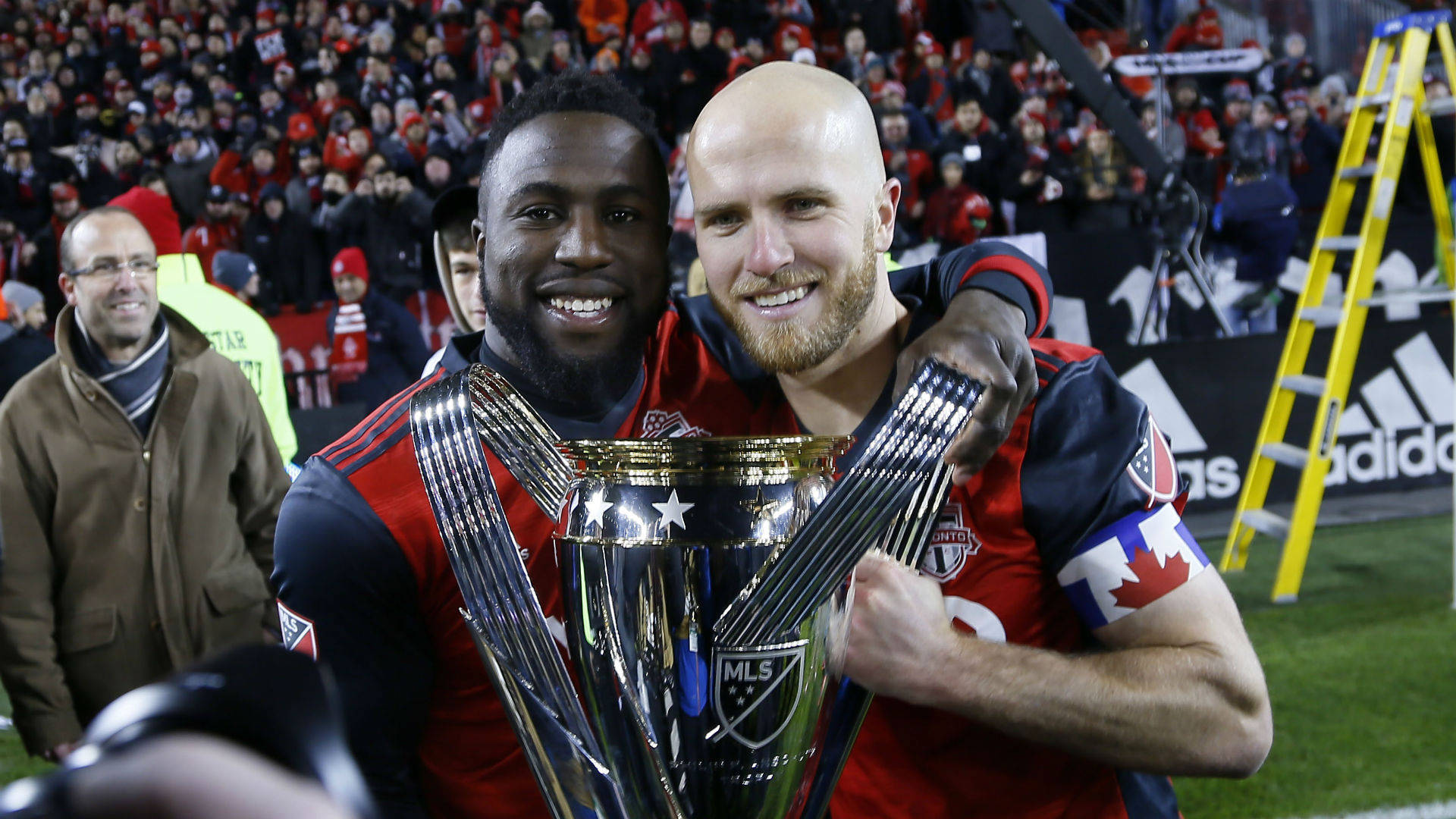足球 桌布 壁紙 — 兩名微笑的足球員在球場上擁抱，手持閃亮的MLS Cup獎盃，背景是歡呼的球迷。