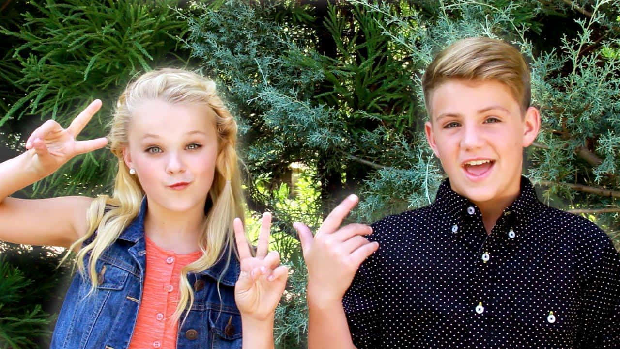 JoJo Siwa 桌布 壁紙 — 兩名微笑的青少年在戶外愉快地擺姿勢，身後有綠色樹葉，做出和平手勢。