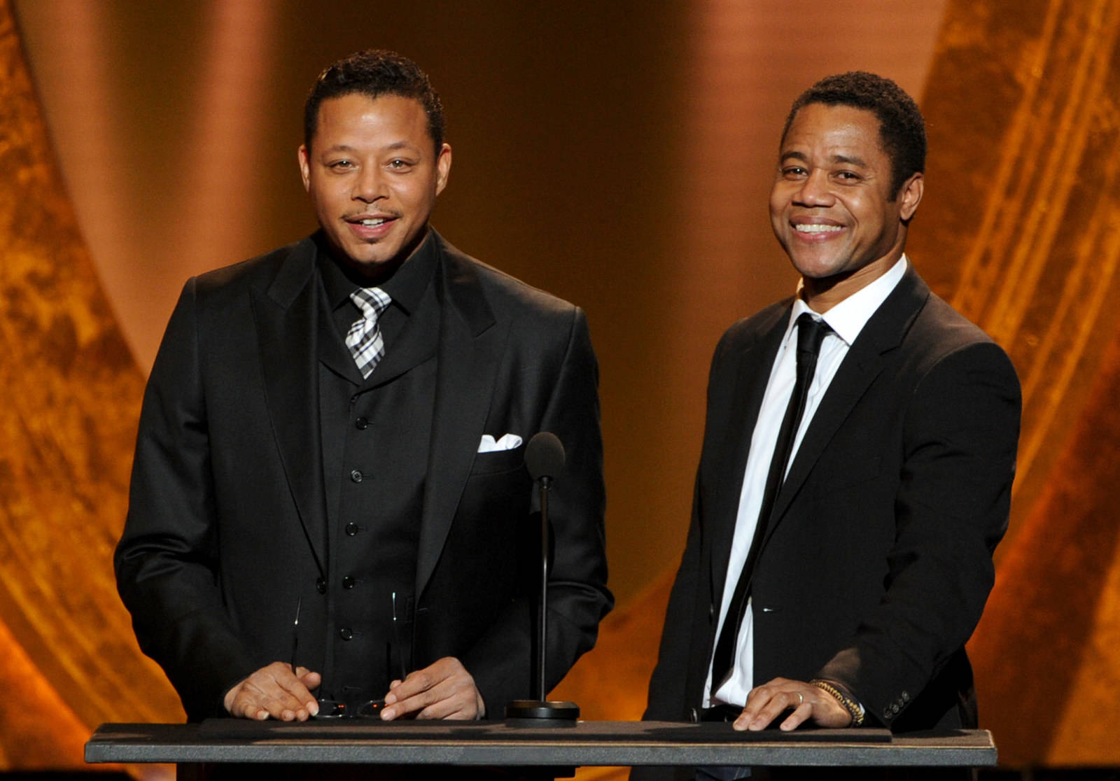 Cuba Gooding Jr 桌布 壁紙 — 兩名微笑的黑西裝男子站在一個溫暖燈光的舞台上的講台後方，背景有金色垂簾。