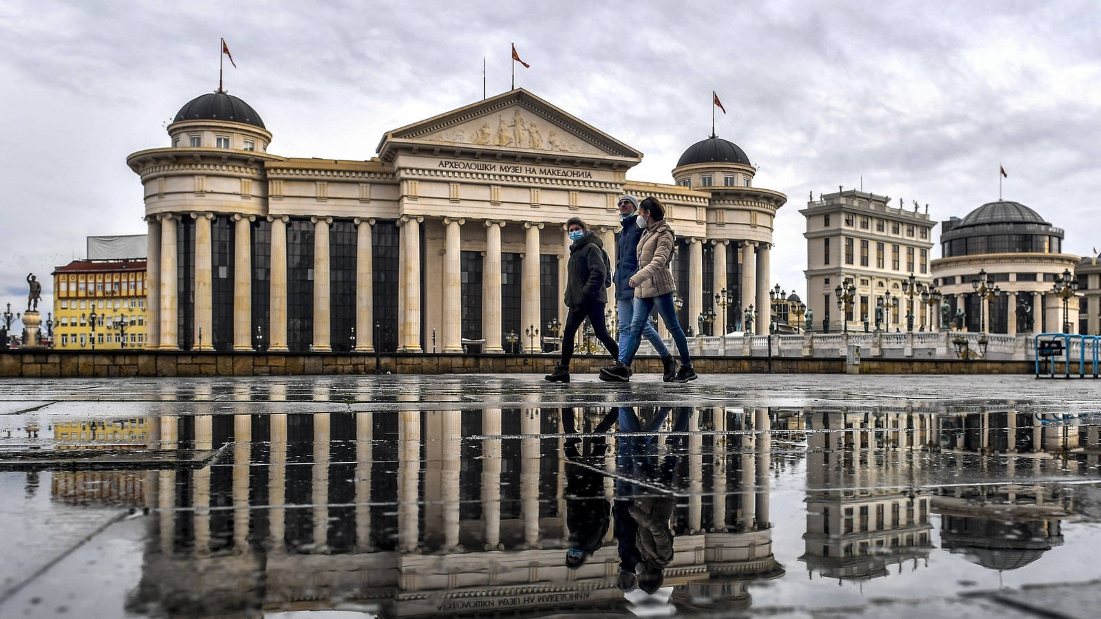 Macedonia 桌布 壁紙 — 兩名戴口罩的人走過一個濕漉漉的廣場，廣場反射出一座宏偉的新古典主義建築，天空多雲。