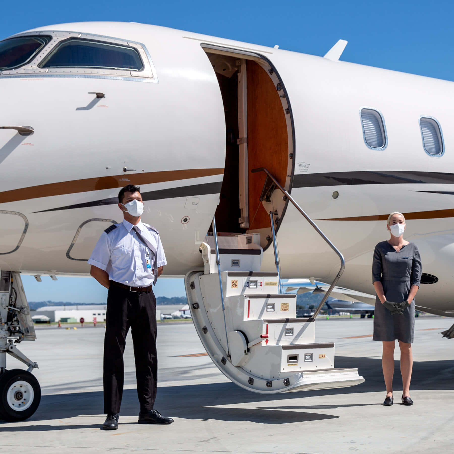 flying 桌布 壁紙 — 兩名戴口罩的空中服務員站在私人飛機敞開的艙門旁，背景是晴朗的藍天和陽光普照的停機坪。