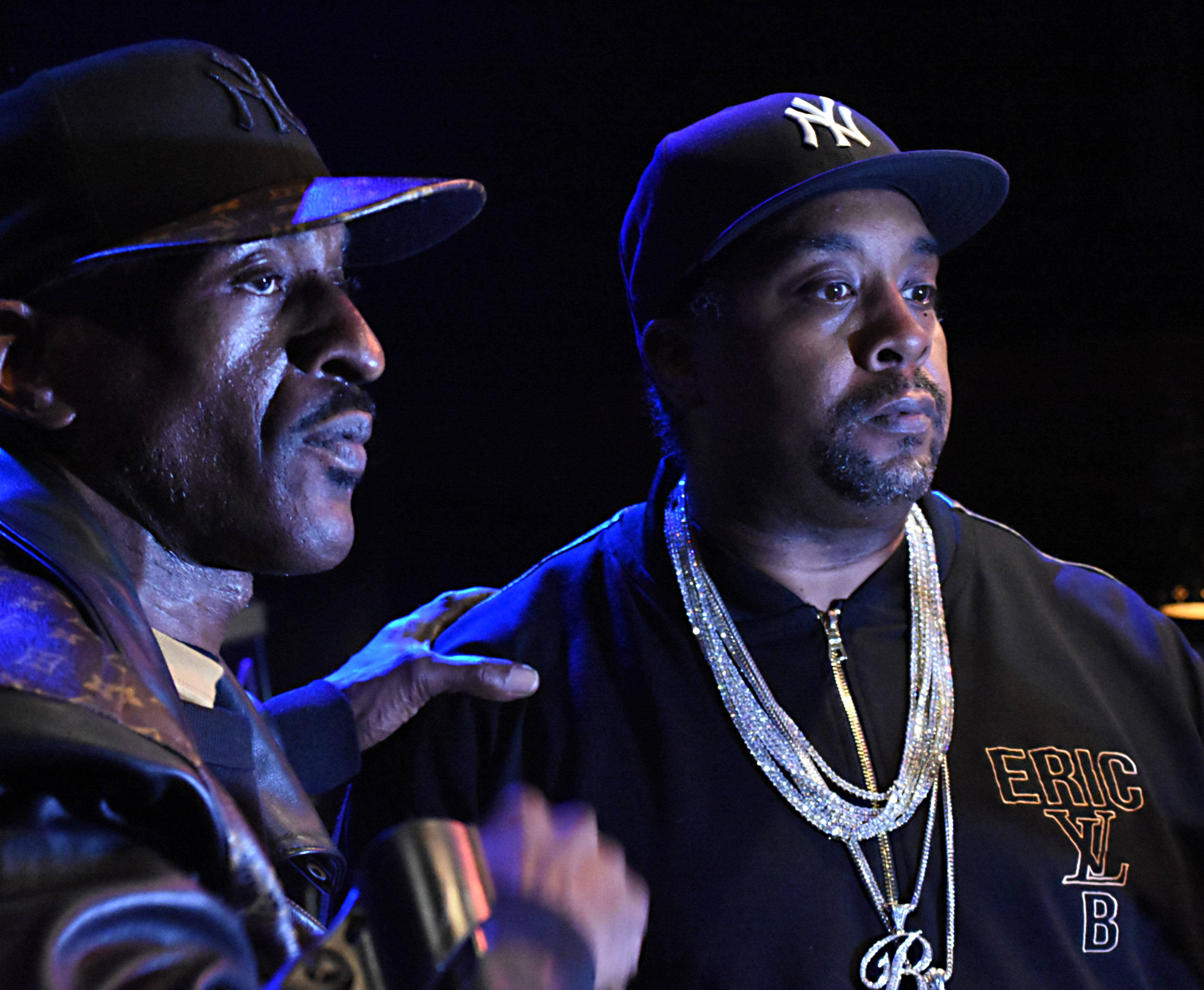Rappers 桌布 壁紙 — 兩名戴帽子和鏈條的男子站在藍色舞台燈光下，其中一人向另一人做手勢，背景為深色。