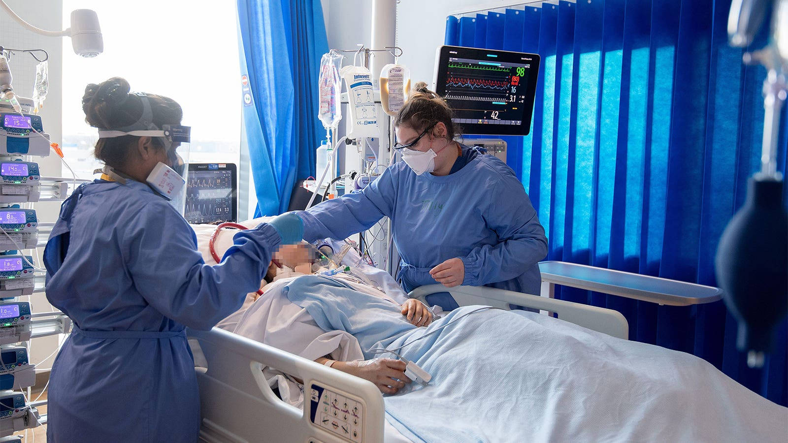 Hospital 桌布 壁紙 — 兩名戴著口罩、身穿藍色手術服的護士正在照顧一名躺在醫院病床上的病人，周圍是醫療監視器和靜脈注射設備。