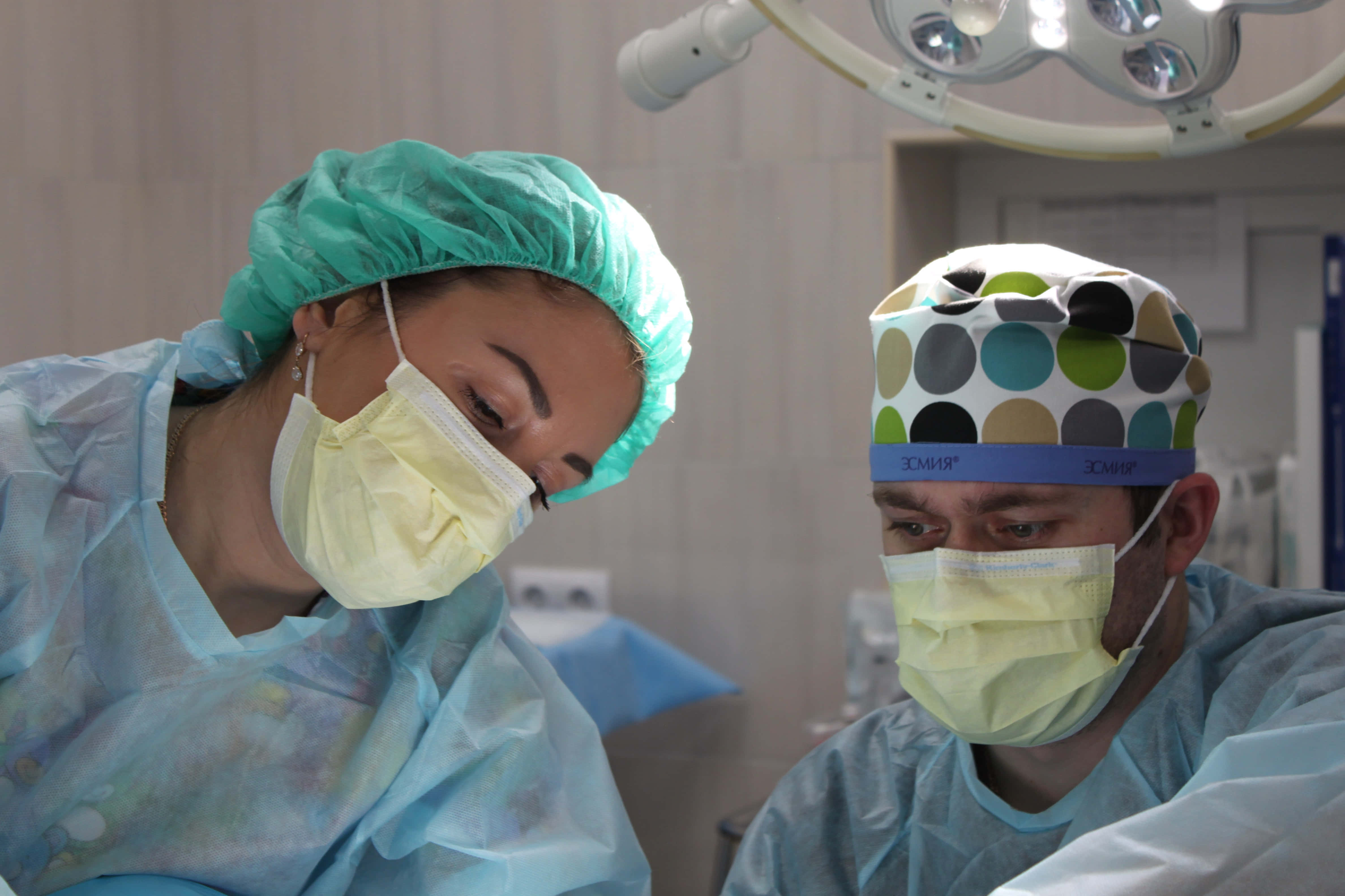 Hospital 桌布 壁紙 — 兩名戴著口罩和帽子的外科醫生在明亮的手術燈下專注地進行手術。