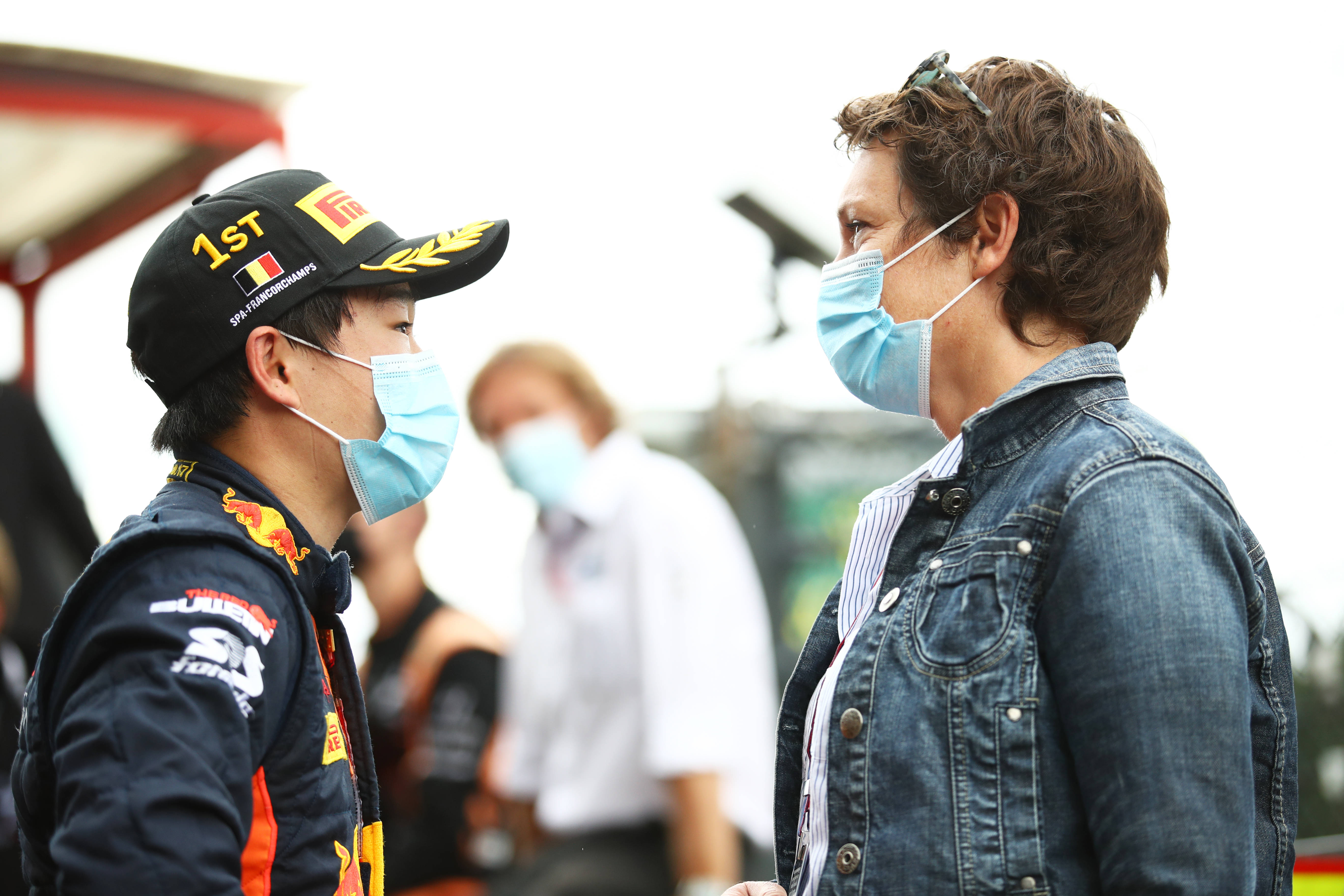 F1 桌布 壁紙 — 兩名戴著口罩的人在賽車活動中交談，一人穿著Red Bull賽車裝備，另一人穿著牛仔外套。