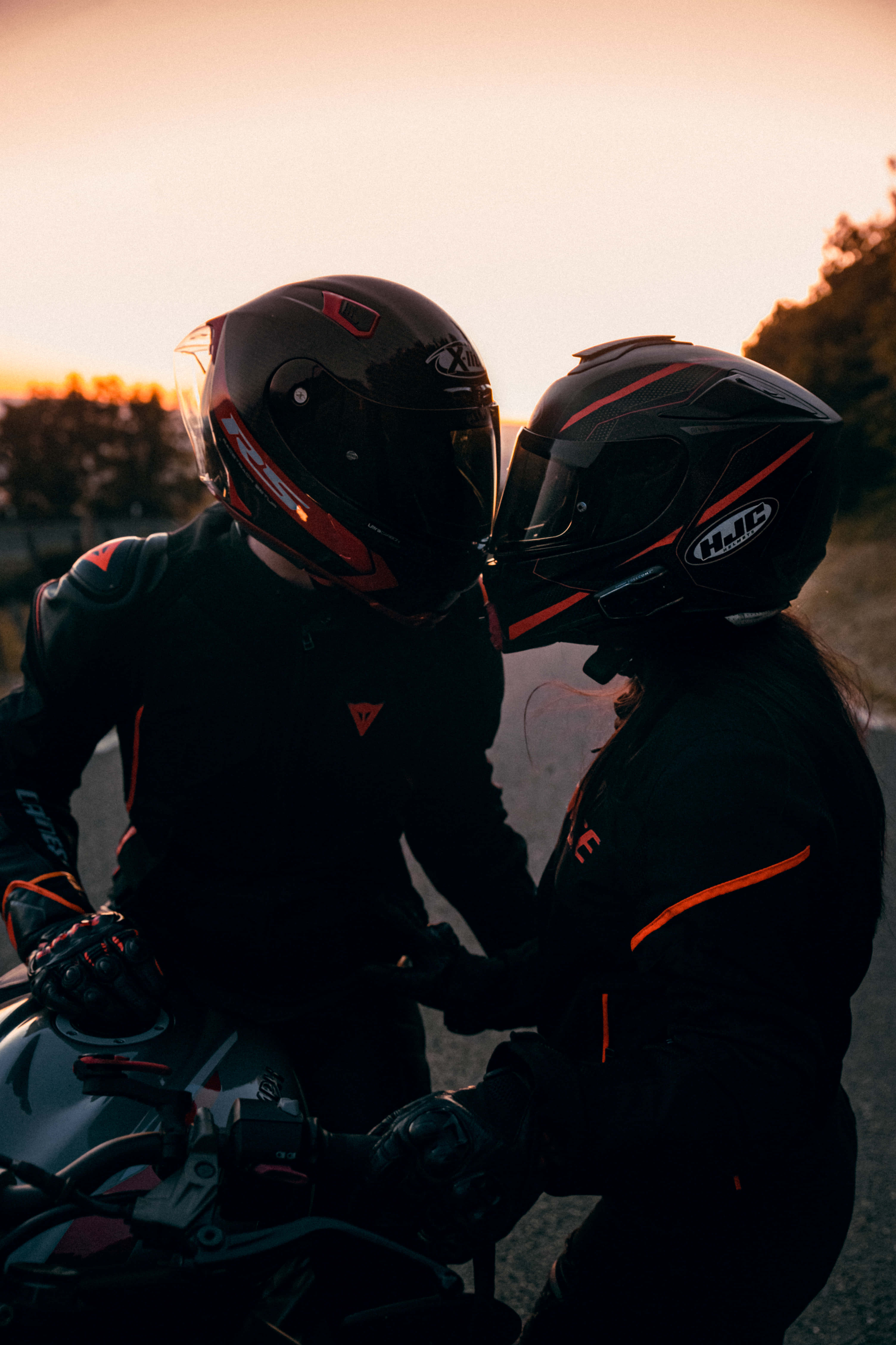 biker 桌布 壁紙 — 兩名戴著帶有橙色點綴的黑色頭盔的摩托車手，在日落時分的路上分享親密時刻。