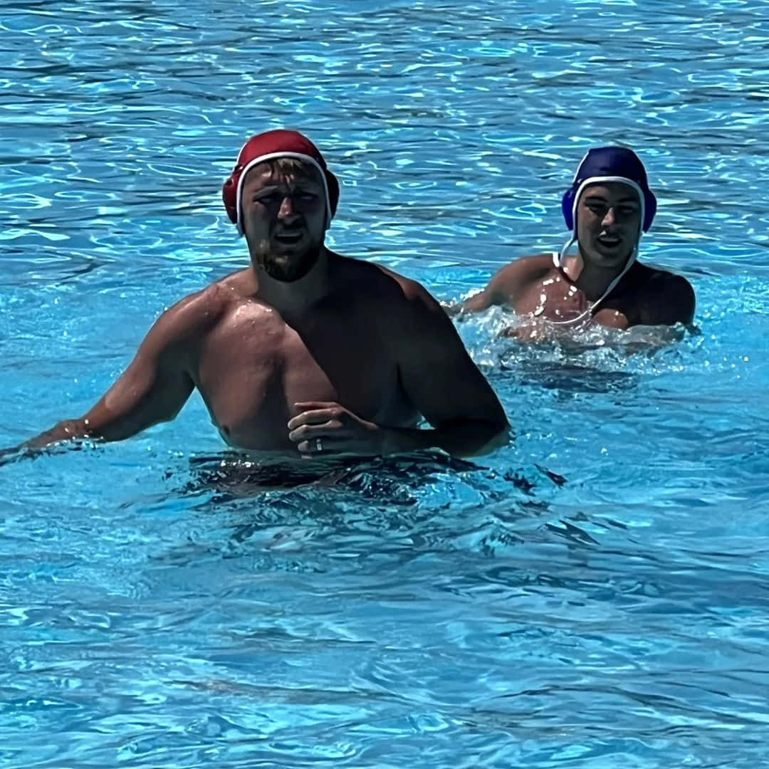 Swimming 桌布 壁紙 — 兩名戴著紅色和藍色泳帽的水球運動員在晴朗的天空下，在明亮的藍色泳池水中激烈競爭。
