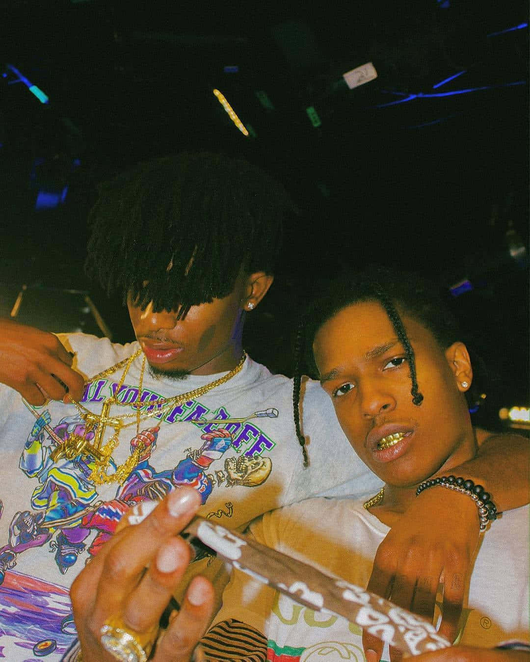 Playboi Carti HD 桌布 壁紙 — 兩名戴金牙和鏈條的年輕男子在燈光昏暗、霓虹燈閃爍的俱樂部中激烈擺姿勢。