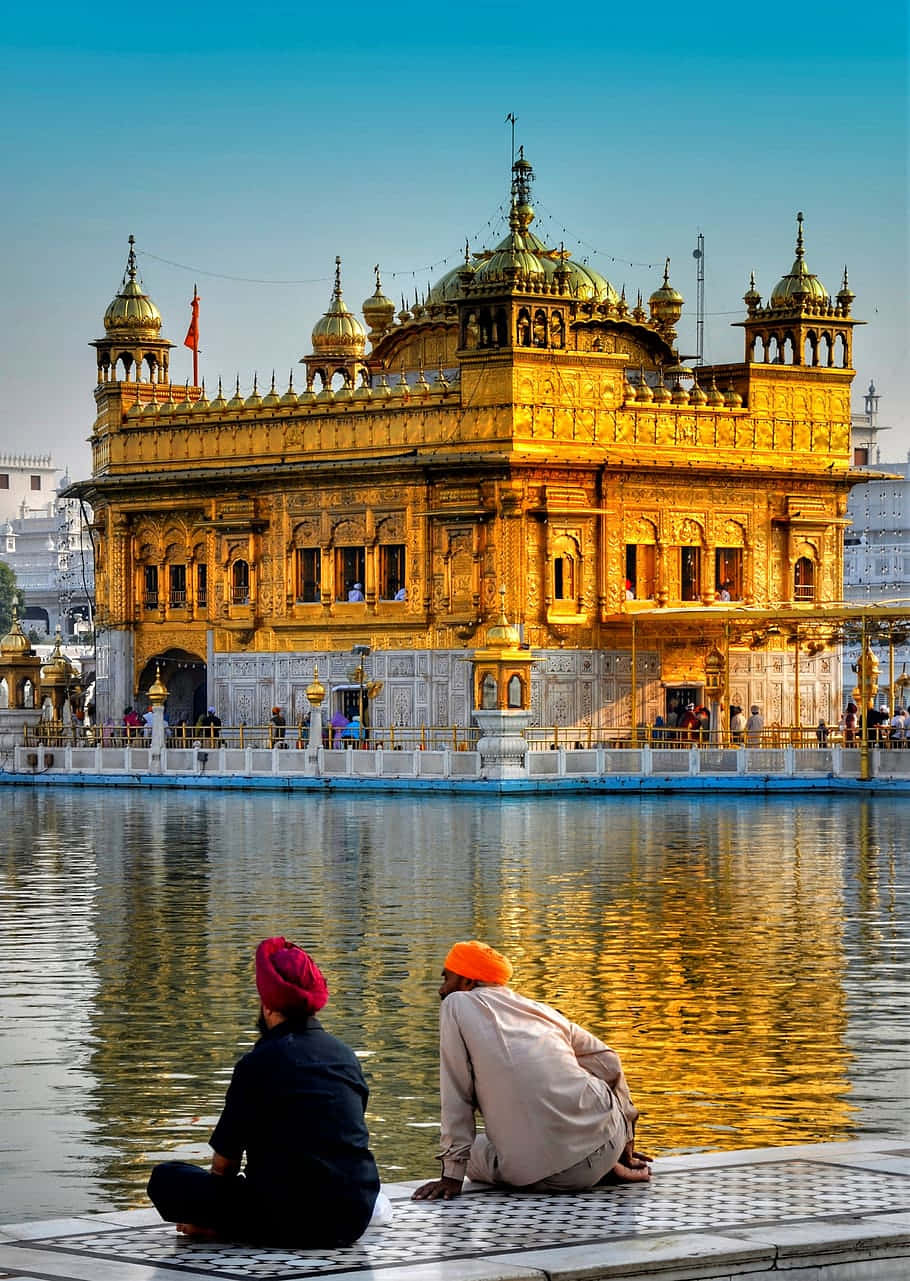 Golden Temple 桌布 壁紙 — 兩名戴頭巾的男子平靜地坐在水邊，水面倒映著金色哈曼迪爾薩希布神廟，天空湛藍清澈。