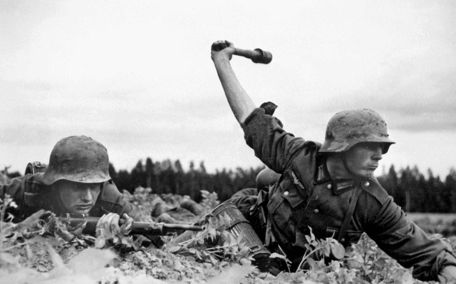 Ww2 桌布 壁紙 — 兩名戴頭盔的士兵蹲在泥濘的地形中，一人舉起手榴彈，另一人在多雲的天空下瞄準步槍。