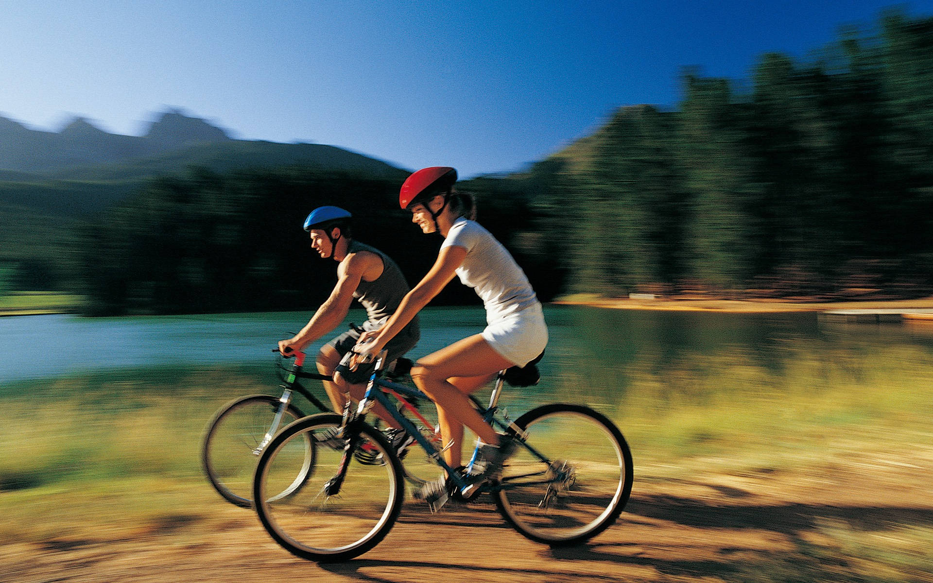 bike riding 桌布 壁紙 — 兩名戴頭盔的自行車騎士在陽光普照的小徑上並排騎行，旁邊是湖泊，背景有模糊的綠色山丘和藍天在移動。