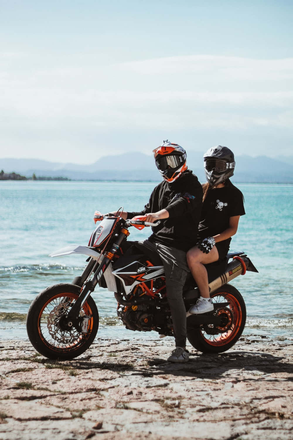 biker 桌布 壁紙 — 兩名戴頭盔的騎士坐在一輛KTM越野機車上，停泊在岩石海灘，背景是碧綠海水、遠山和明亮的天空。