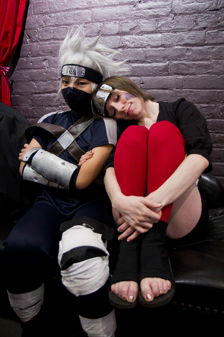 Kakashi And Rin 桌布 壁紙 — 兩名扮演火影忍者角色的Cosplayer緊貼著坐在磚牆前的皮沙發上，一人將頭靠在另一人的肩膀上。