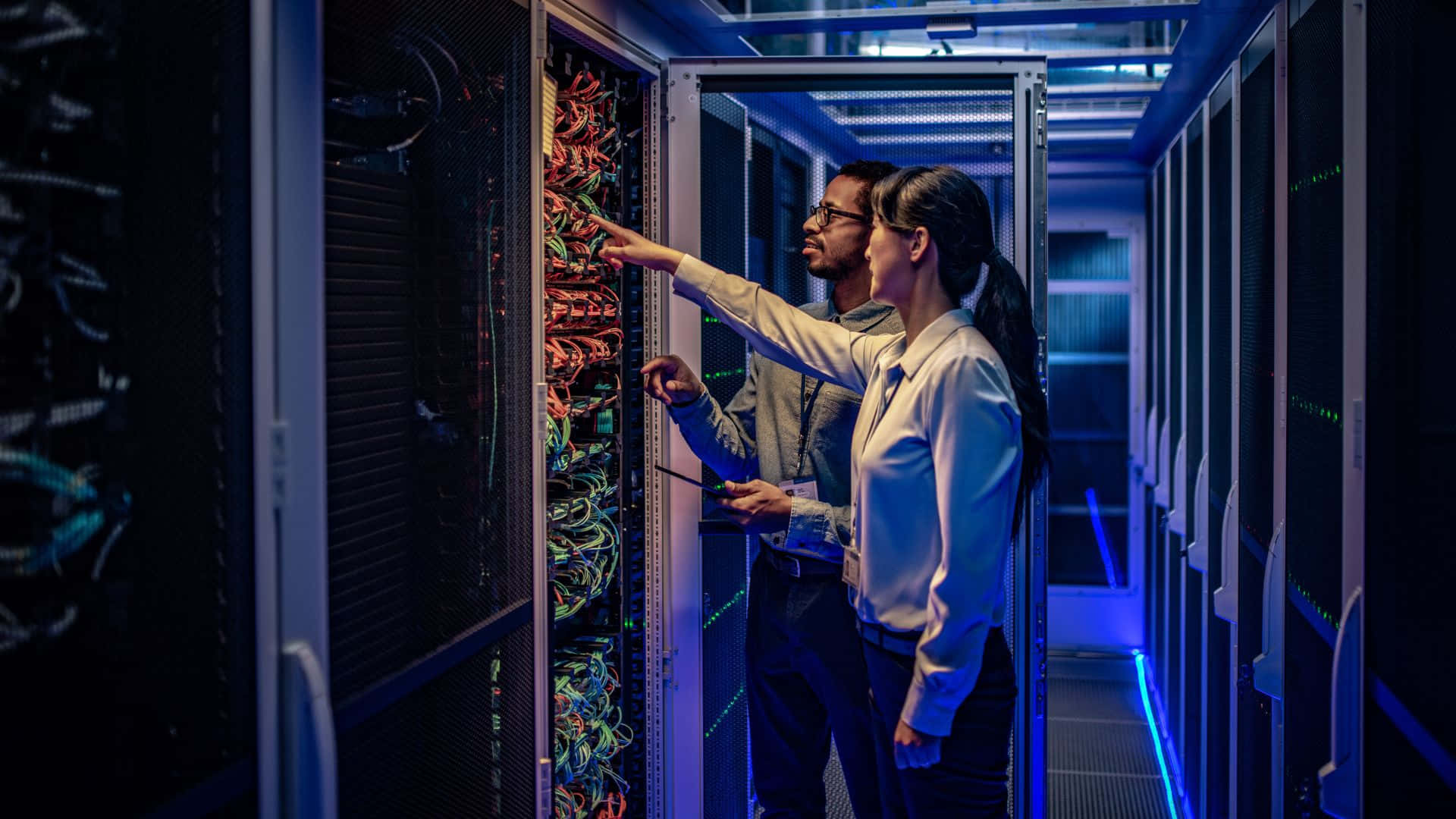 Gaming Servers 桌布 壁紙 — 兩名技術人員在昏暗的資料中心檢查發光的伺服器機架，沐浴在藍光中。
