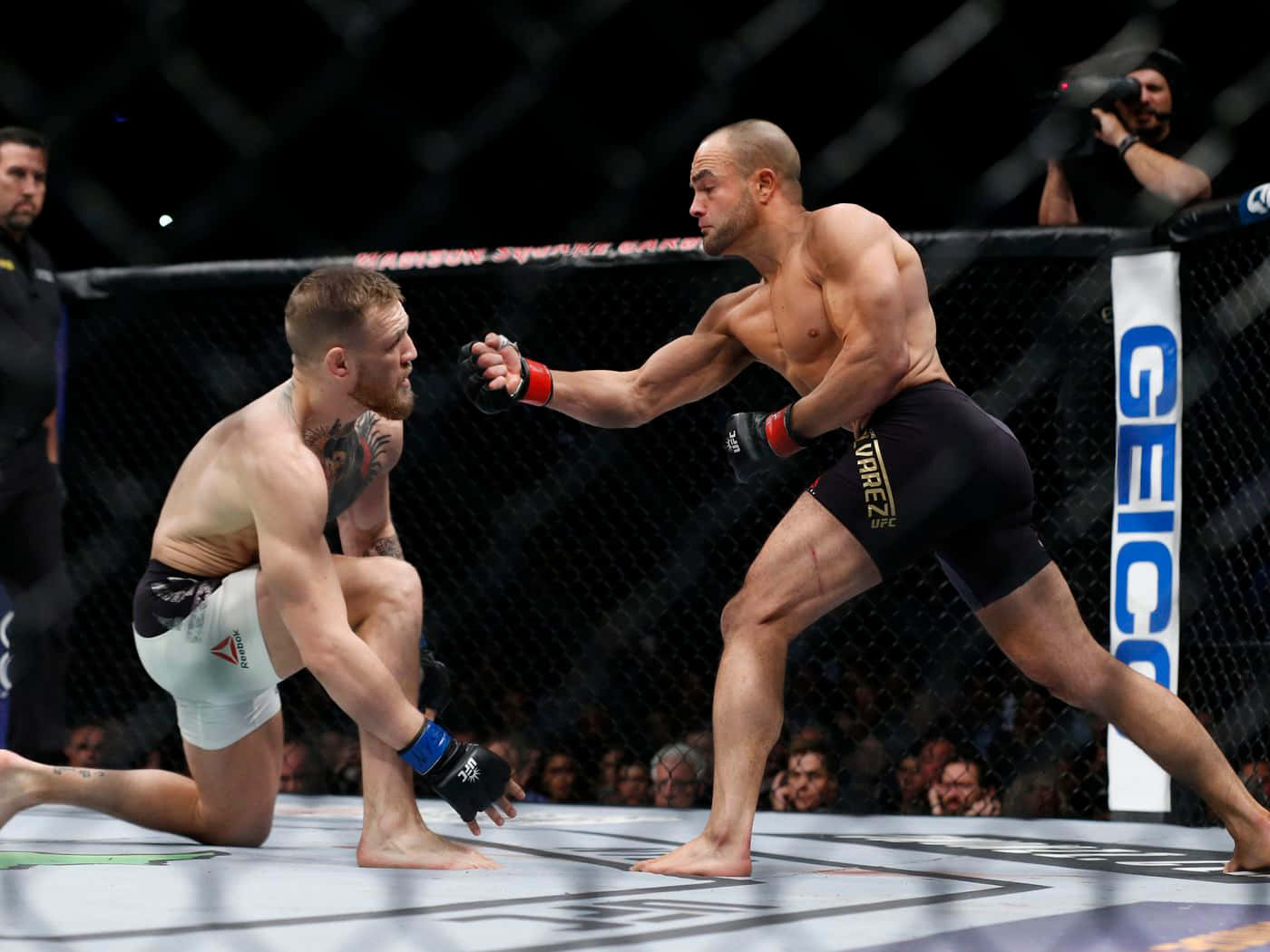 boxer 桌布 壁紙 — 兩名拳手在UFC八角籠中交鋒，一人向前猛衝出拳，另一人在明亮的場館燈光下防守性地跪地。