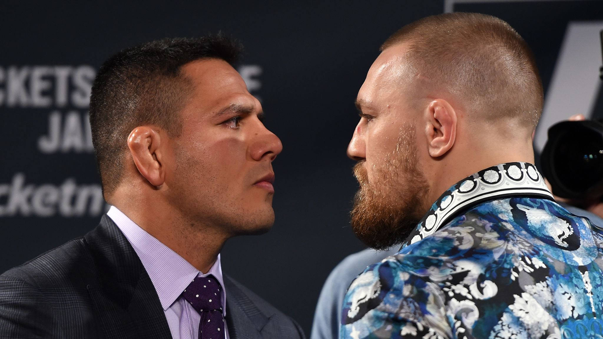 UFC 桌布 壁紙 — 兩名拳手在記者會上緊張對峙，一人身穿西裝，另一人則穿著大膽的藍色圖案外套。