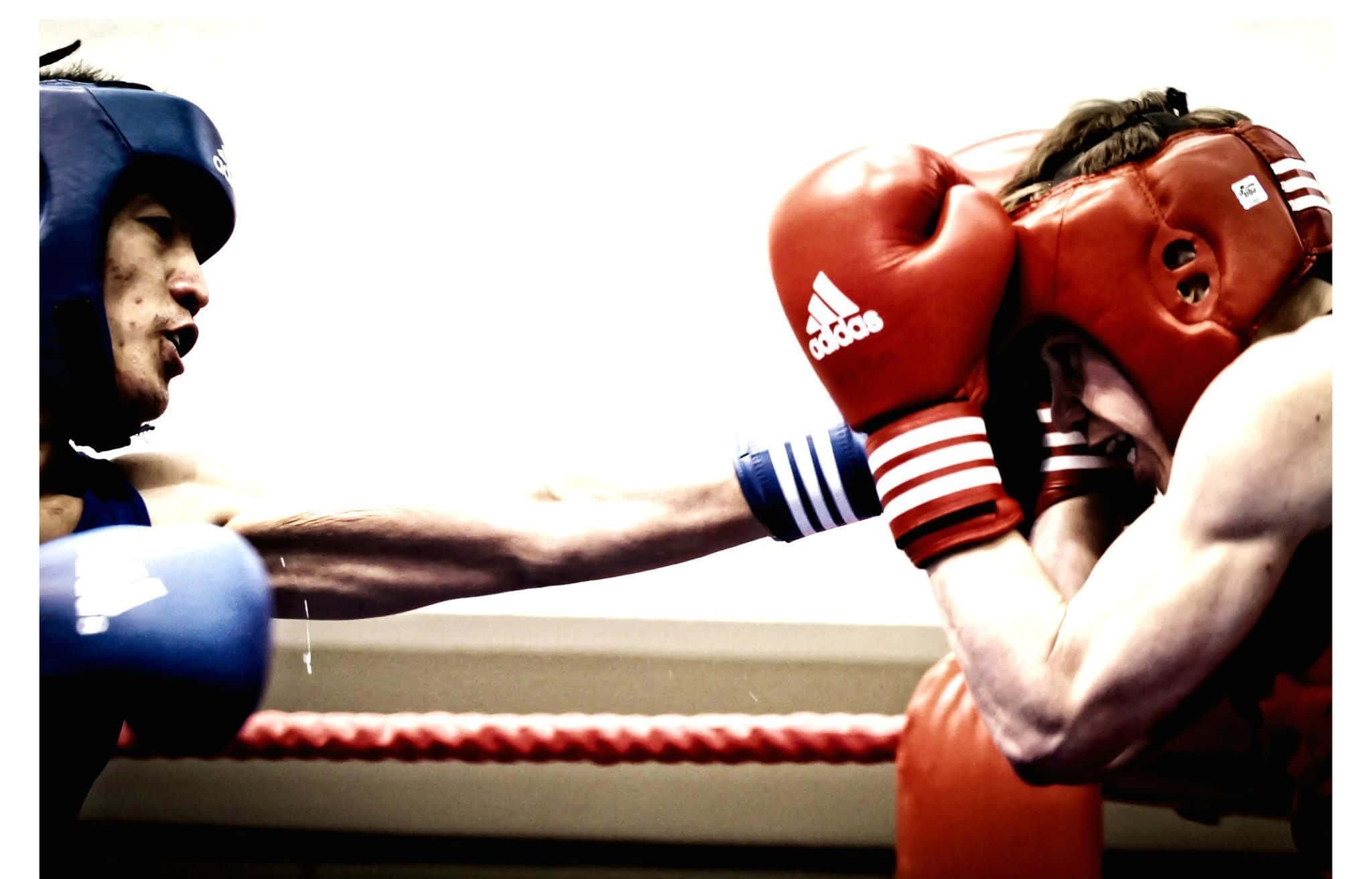 Boxing 桌布 壁紙 — 兩名拳擊手在擂台上交鋒，一人出拳，另一人則以紅色手套和頭盔防守。