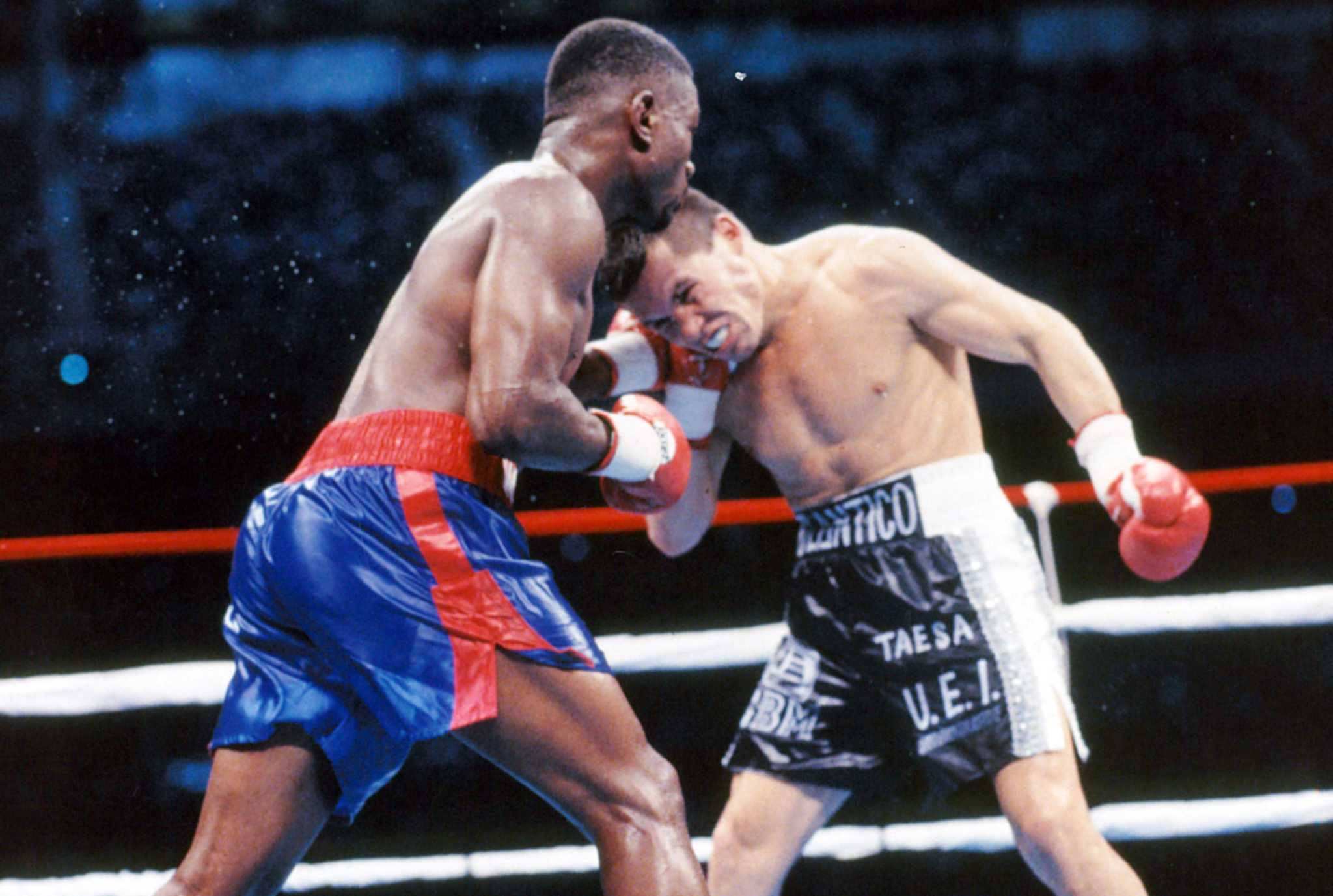 Boxing 桌布 壁紙 — 兩名拳擊手在激烈的擂台比賽中交鋒，其中一人在明亮的場館燈光下向對手的臉部猛烈出拳。