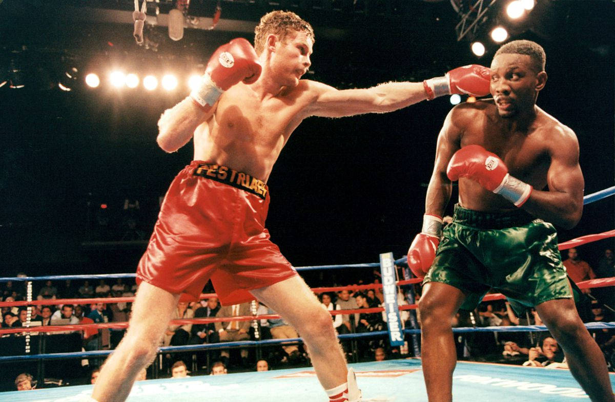 Boxing 桌布 壁紙 — 兩名拳擊手在燈光明亮的擂台上交鋒，一名穿紅色短褲的拳手向穿綠色短褲的對手揮拳。