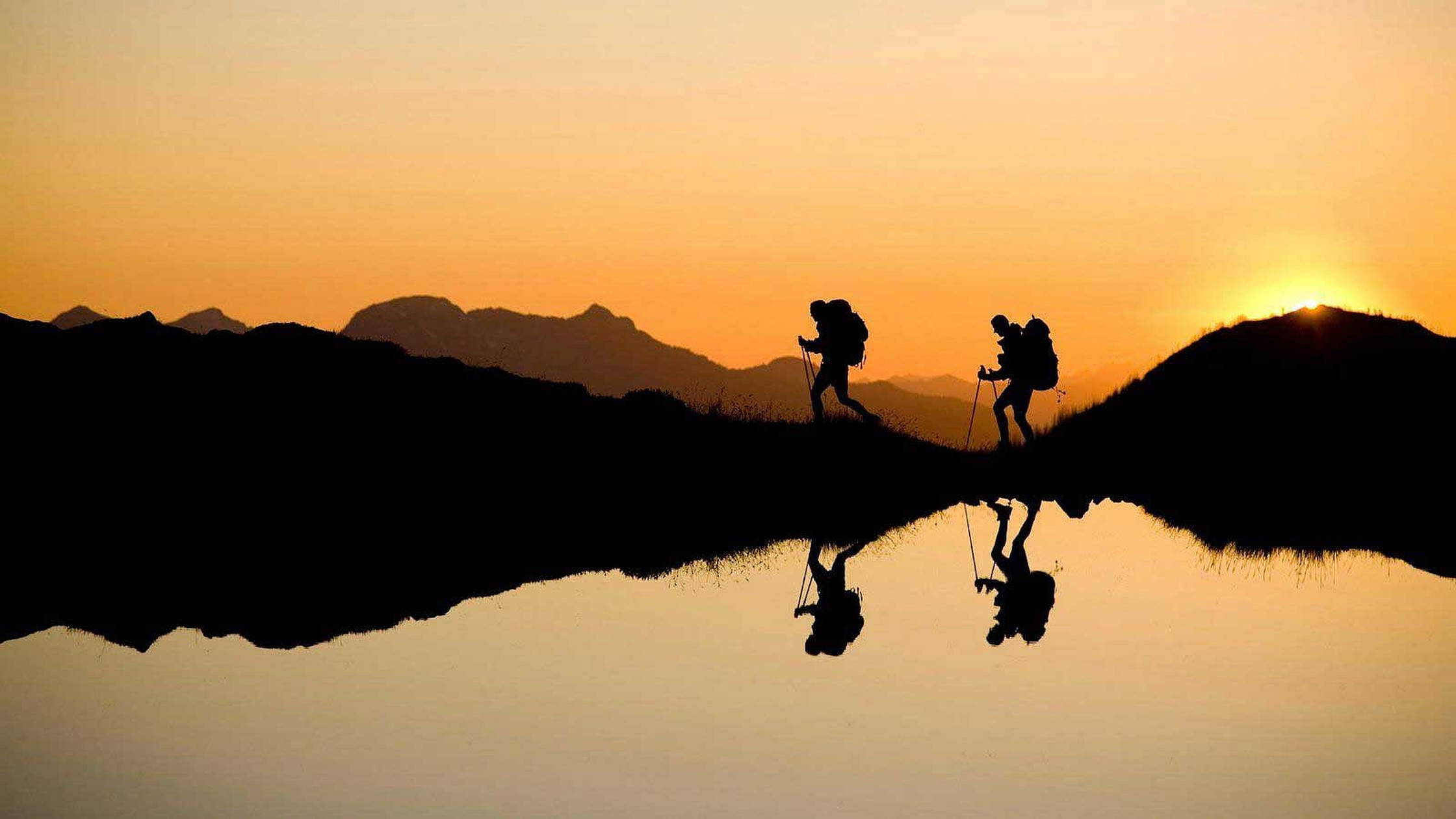 Hiking 桌布 壁紙 — 兩名揹著背包、手持登山杖的遠足者在日落時分穿越草叢山脊，他們的剪影完美倒映在下方平靜的湖面上。