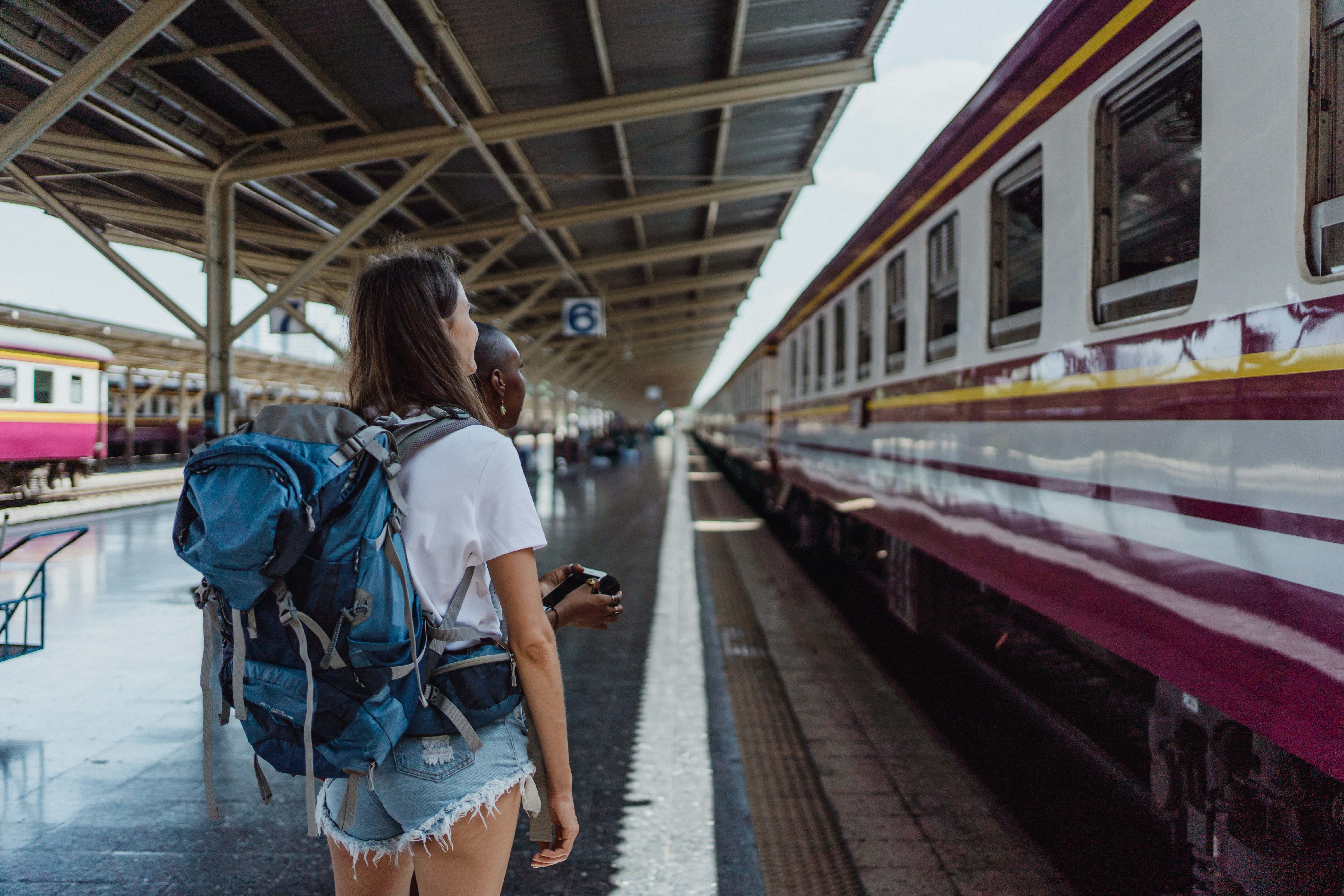 Waiting 桌布 壁紙 — 兩名揹著背包的旅客站在火車月台上，看著一輛栗色和白色的火車在金屬屋頂下抵達。