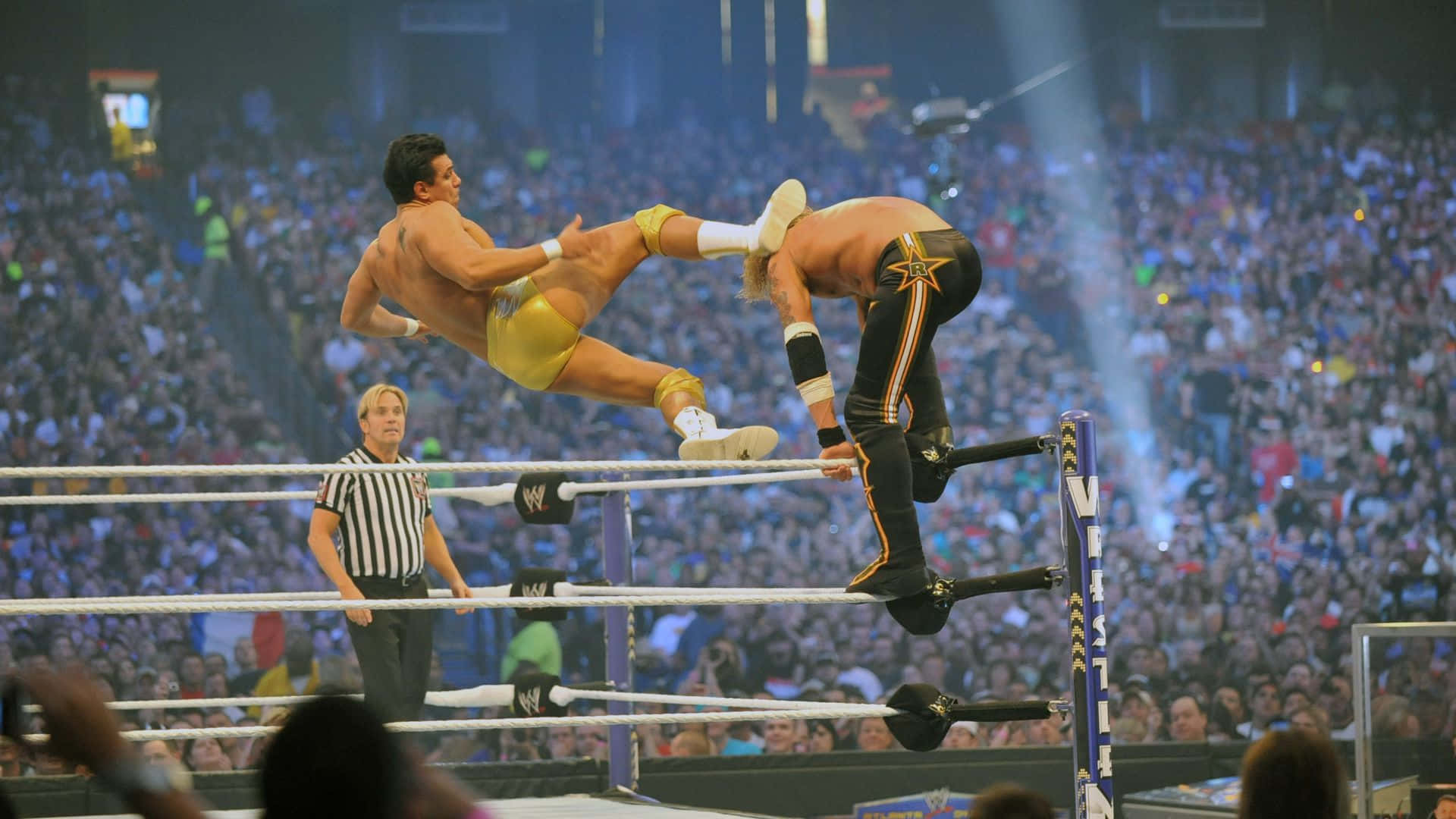 WWE 桌布 壁紙 — 兩名摔角手在半空中進行戲劇性的擂台動作，裁判在旁觀看，下方是座無虛席的場館。