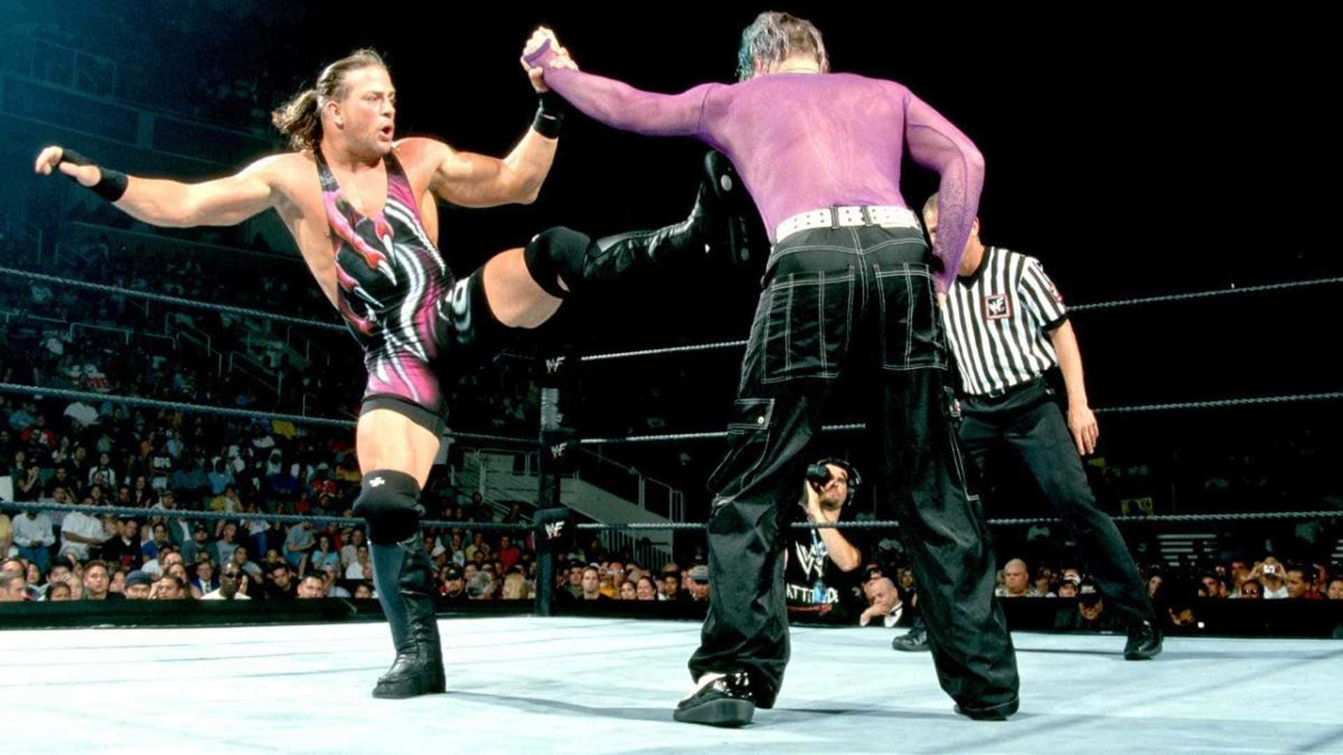 WWE 桌布 壁紙 — 兩名摔角手在擂台上交鋒，其中一人使出高踢，裁判則在座無虛席的場館中監視。