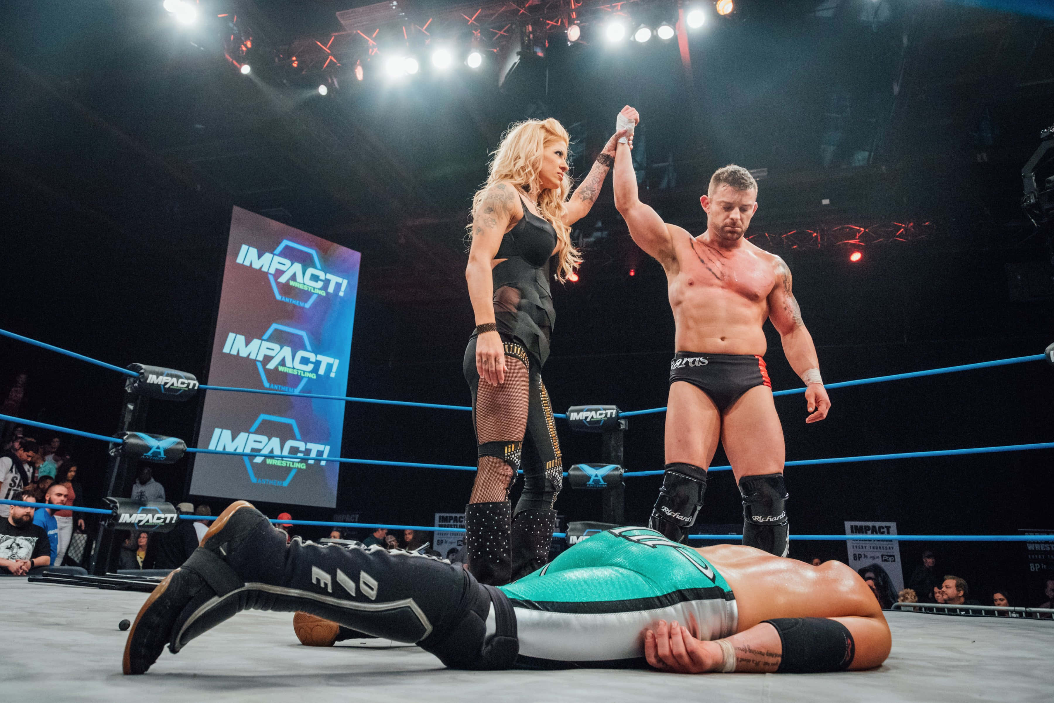 WWE 桌布 壁紙 — 兩名摔角手在擂台上慶祝勝利，而他們被擊敗的對手則躺在明亮的場館燈光下。