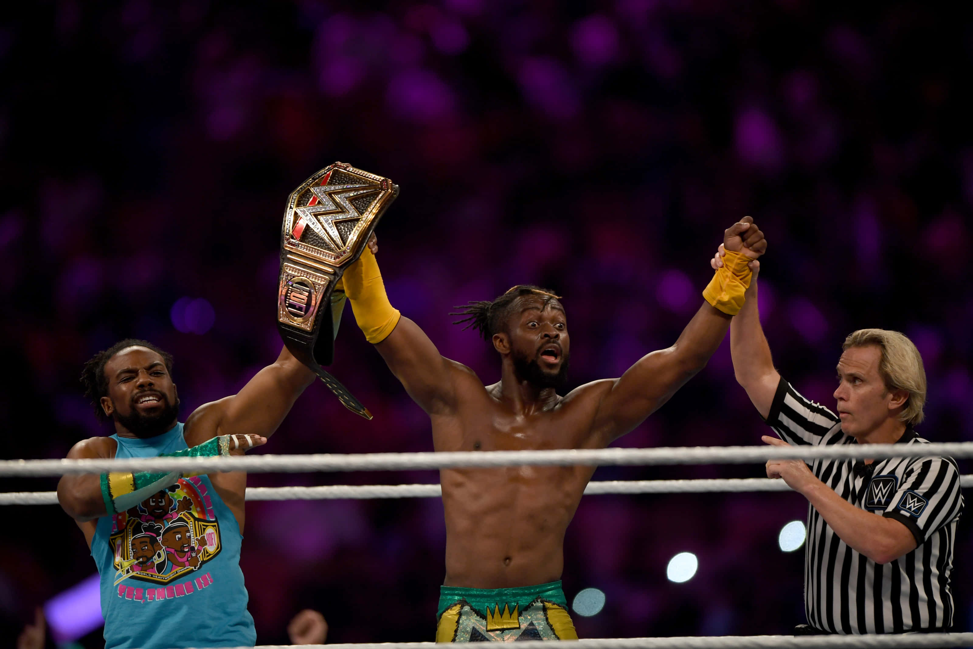 WWE 桌布 壁紙 — 兩名摔角手在擂台上慶祝勝利，裁判在紫色場館燈光下將冠軍腰帶高舉過他們的頭頂。