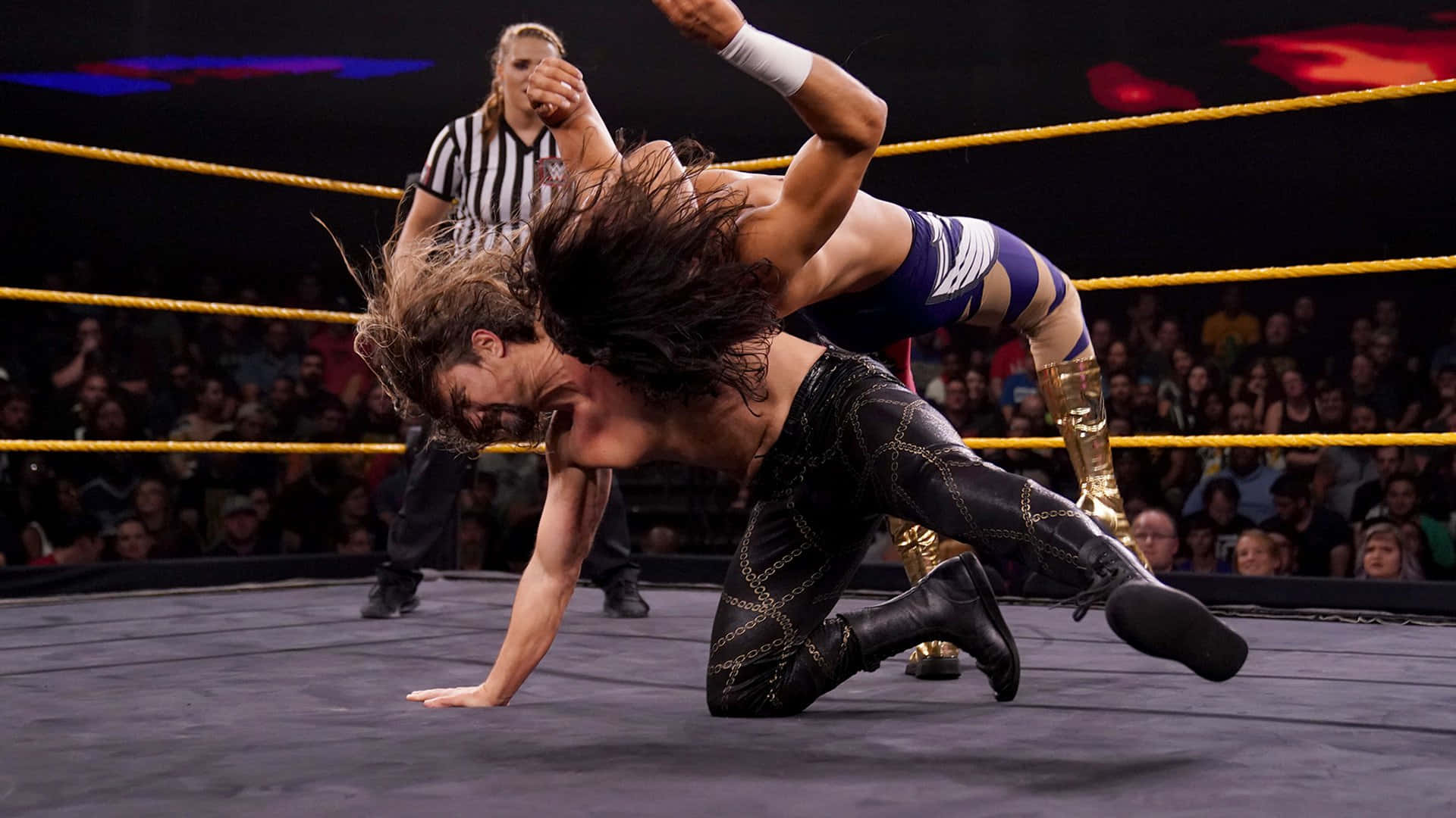 WWE 桌布 壁紙 — 兩名摔角手在擂台上激烈纏鬥，裁判在旁觀看，觀眾在明亮的場館燈光下歡呼。