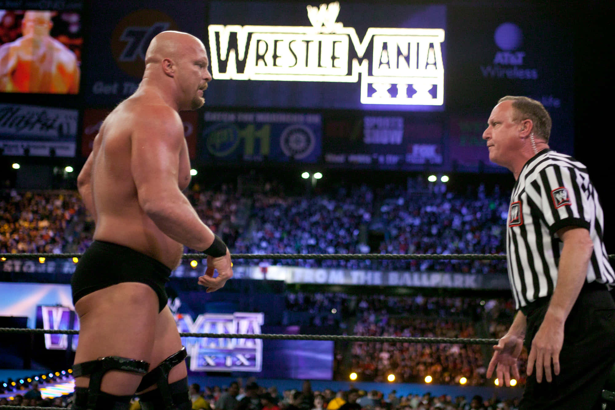 WWE 桌布 壁紙 — 兩名摔角手在明亮的場館燈光下於摔角擂台上對峙，裁判在旁仔細觀察。