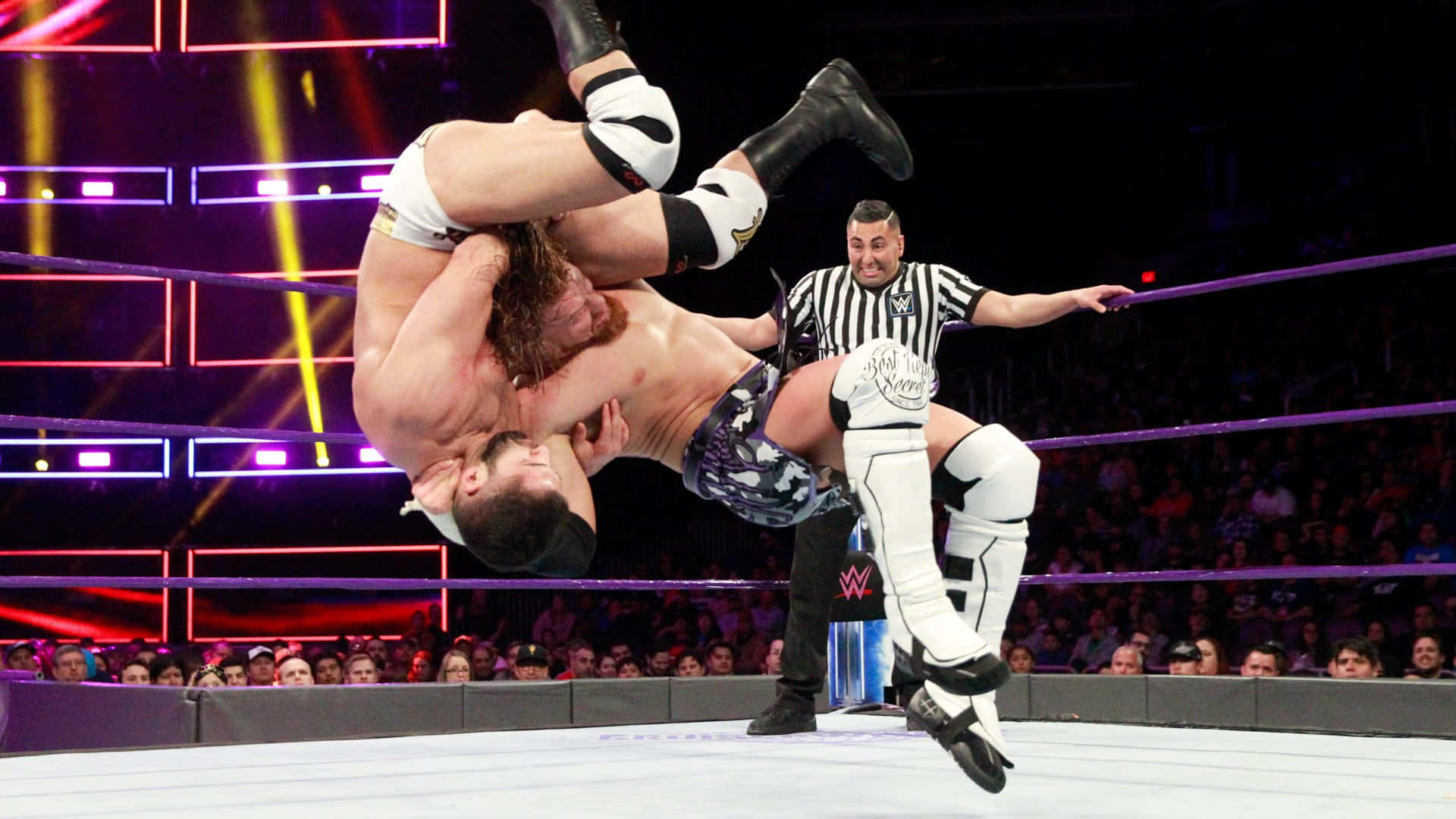 WWE 桌布 壁紙 — 兩名摔角手在燈光明亮的擂台上執行高空飛行動作，裁判觀看，觀眾歡呼。