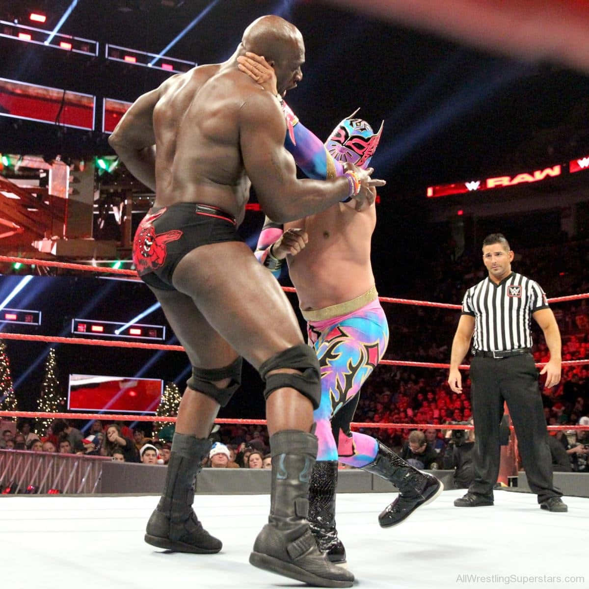 WWE 桌布 壁紙 — 兩名摔角手在燈光明亮的擂台上激烈糾纏，裁判在旁觀看，背景中聖誕樹閃爍著光芒。