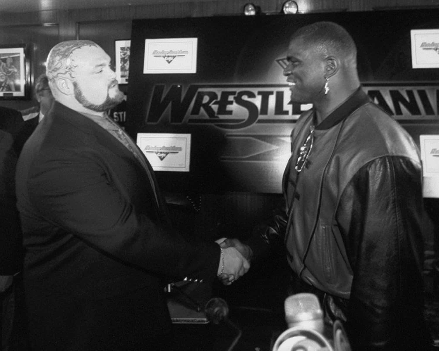WWE 桌布 壁紙 — 兩名摔角手在燈光昏暗的後台區域握手，身後有'WrestleMania'標誌。