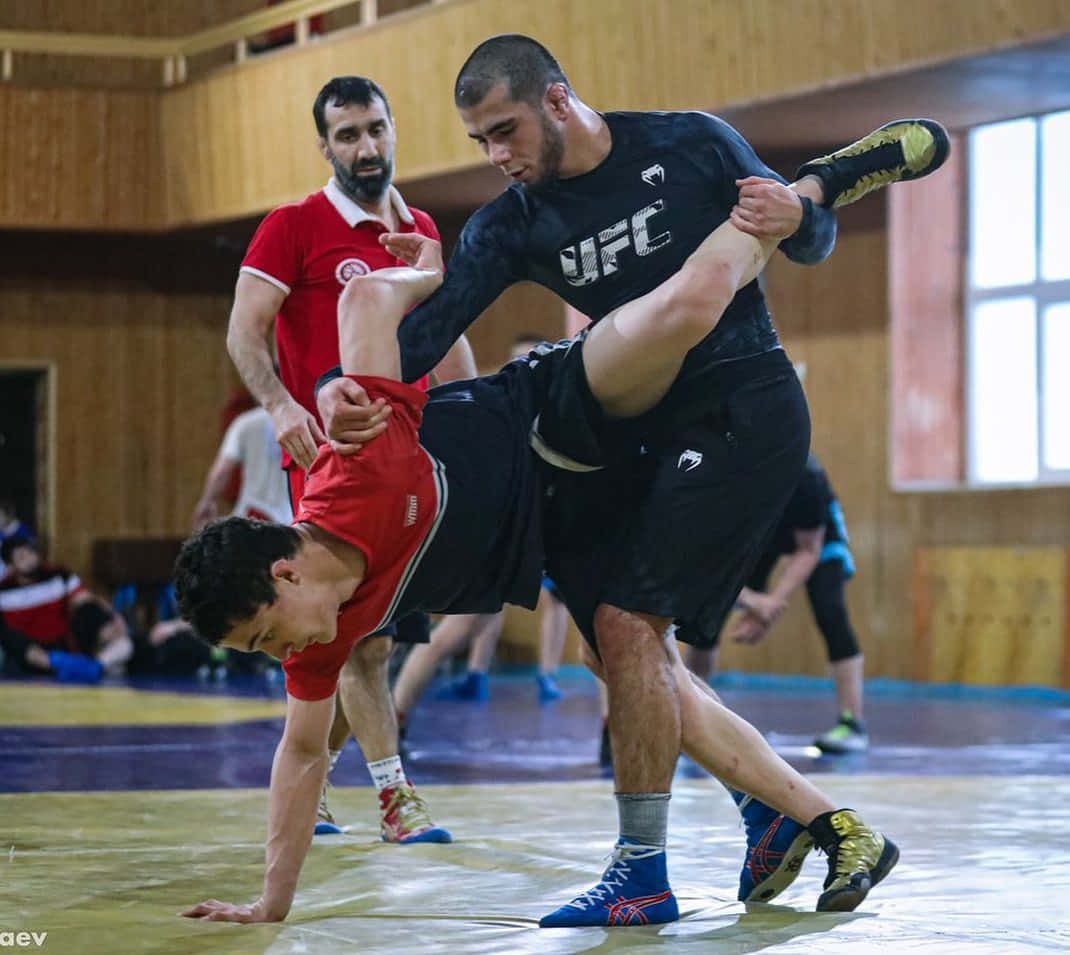 MMA 桌布 壁紙 — 兩名摔跤手在木製室內健身房的墊子上激烈糾纏，觀眾在旁觀看。