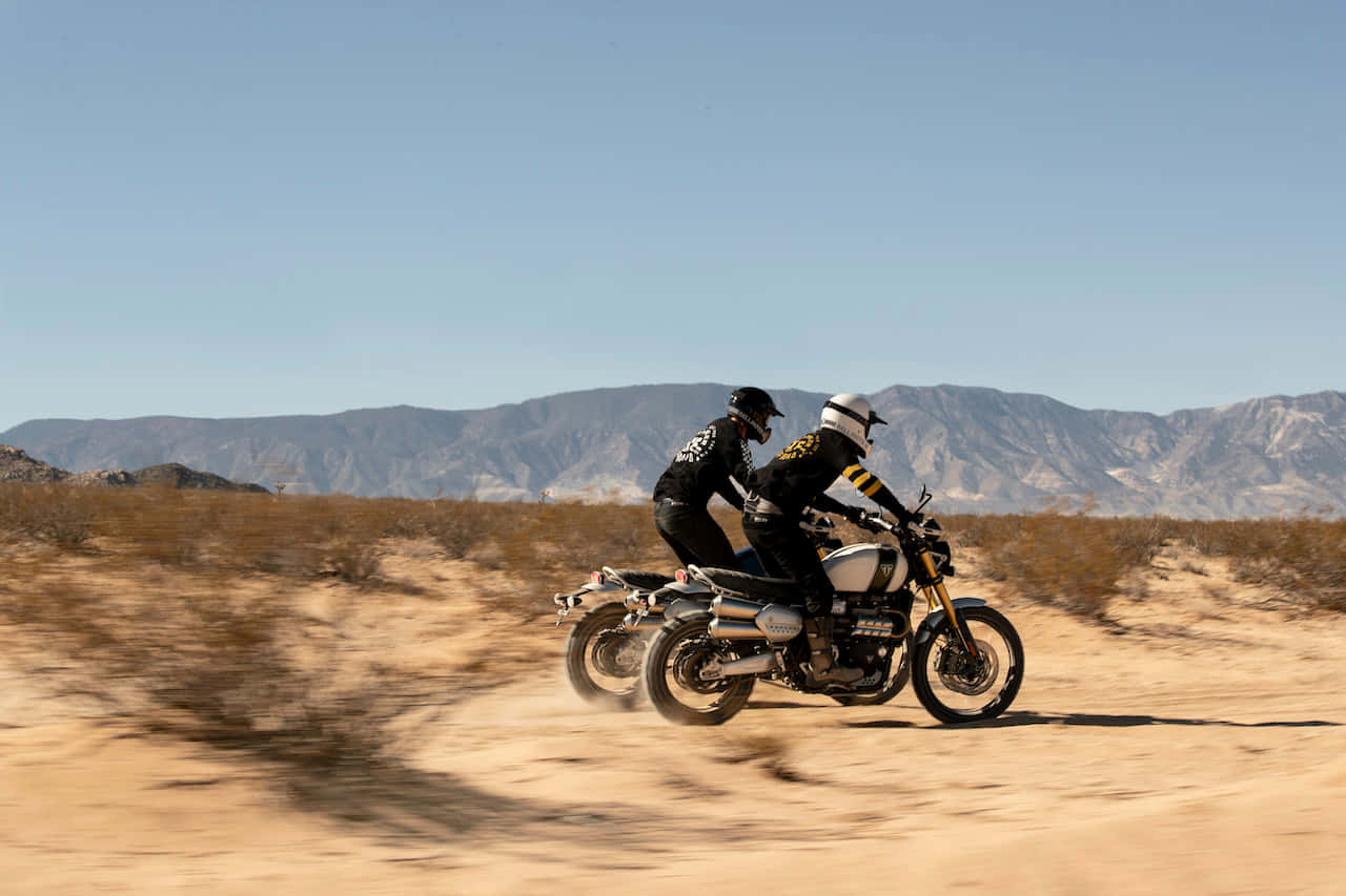 Vehicle 桌布 壁紙 — 兩名摩托車手在烈日曝曬的沙漠上競速，揚起塵土，遠處是崎嶇的山脈，天空清澈湛藍。