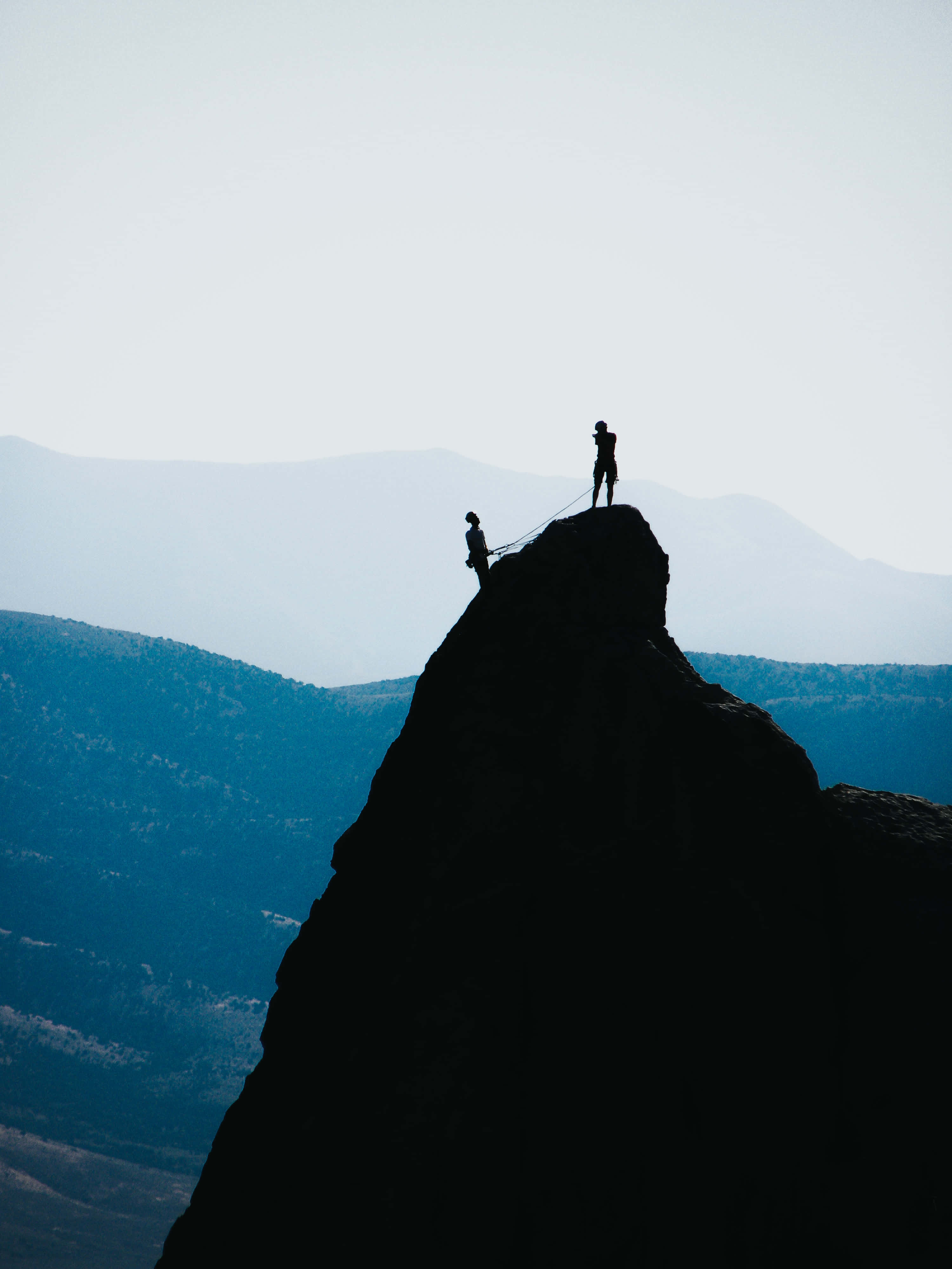 Silhouette 桌布 壁紙 — 兩名攀岩者的剪影站在鋸齒狀山峰上，背景是柔和日光下的朦朧藍色山脈。