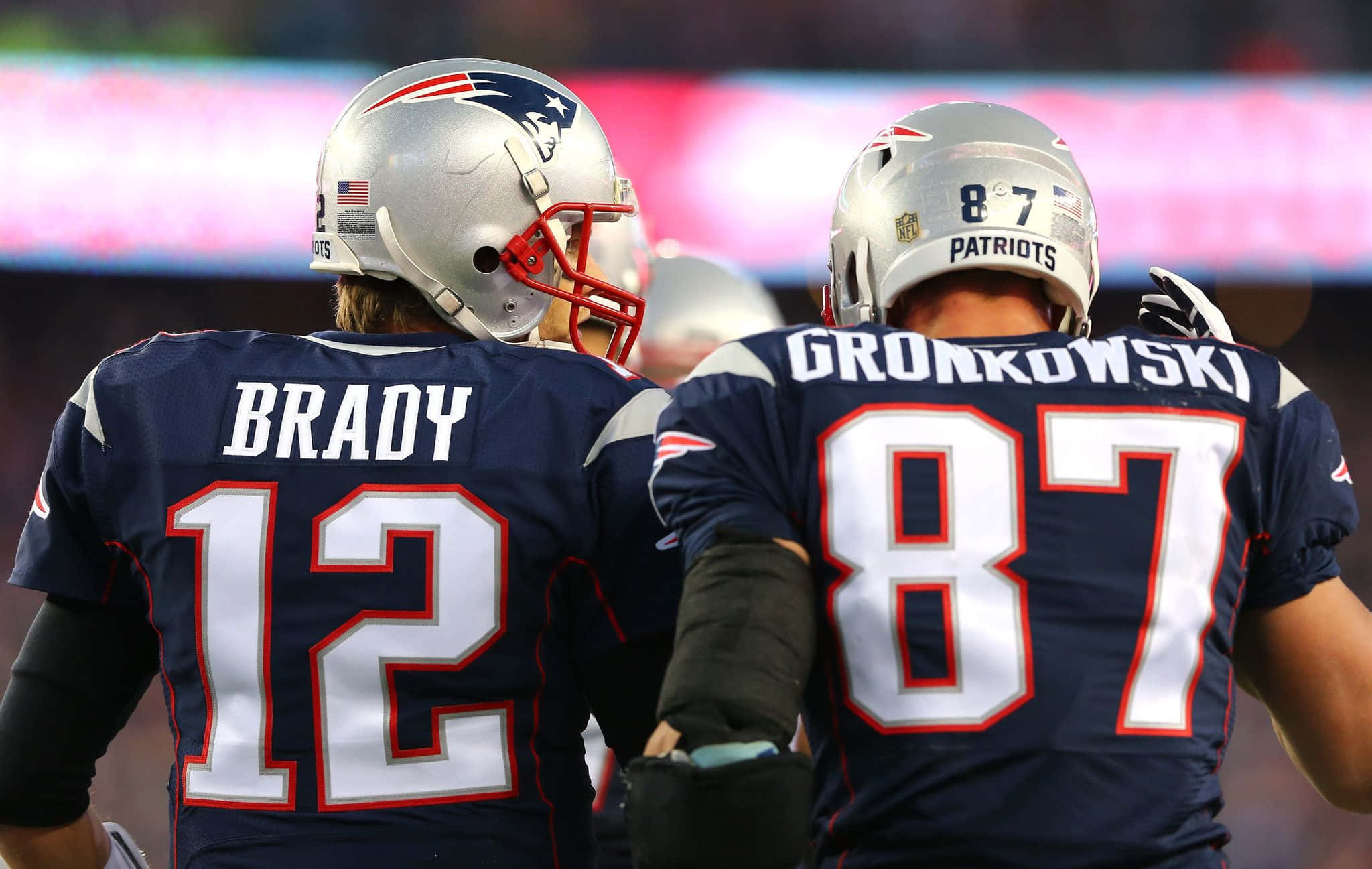 Patriots 桌布 壁紙 — 兩名新英格蘭愛國者隊美式足球員身穿海軍藍球衣，白色號碼12和87，在光線明亮的場地上背對背站立。
